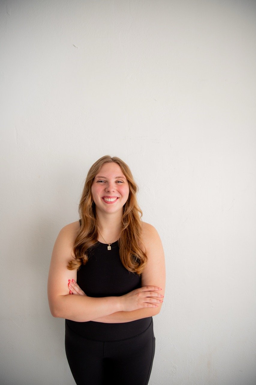 Smiling woman with long, wavy red hair, wearing a black sleeveless top and black pants, standing with arms crossed against a plain white wall.
