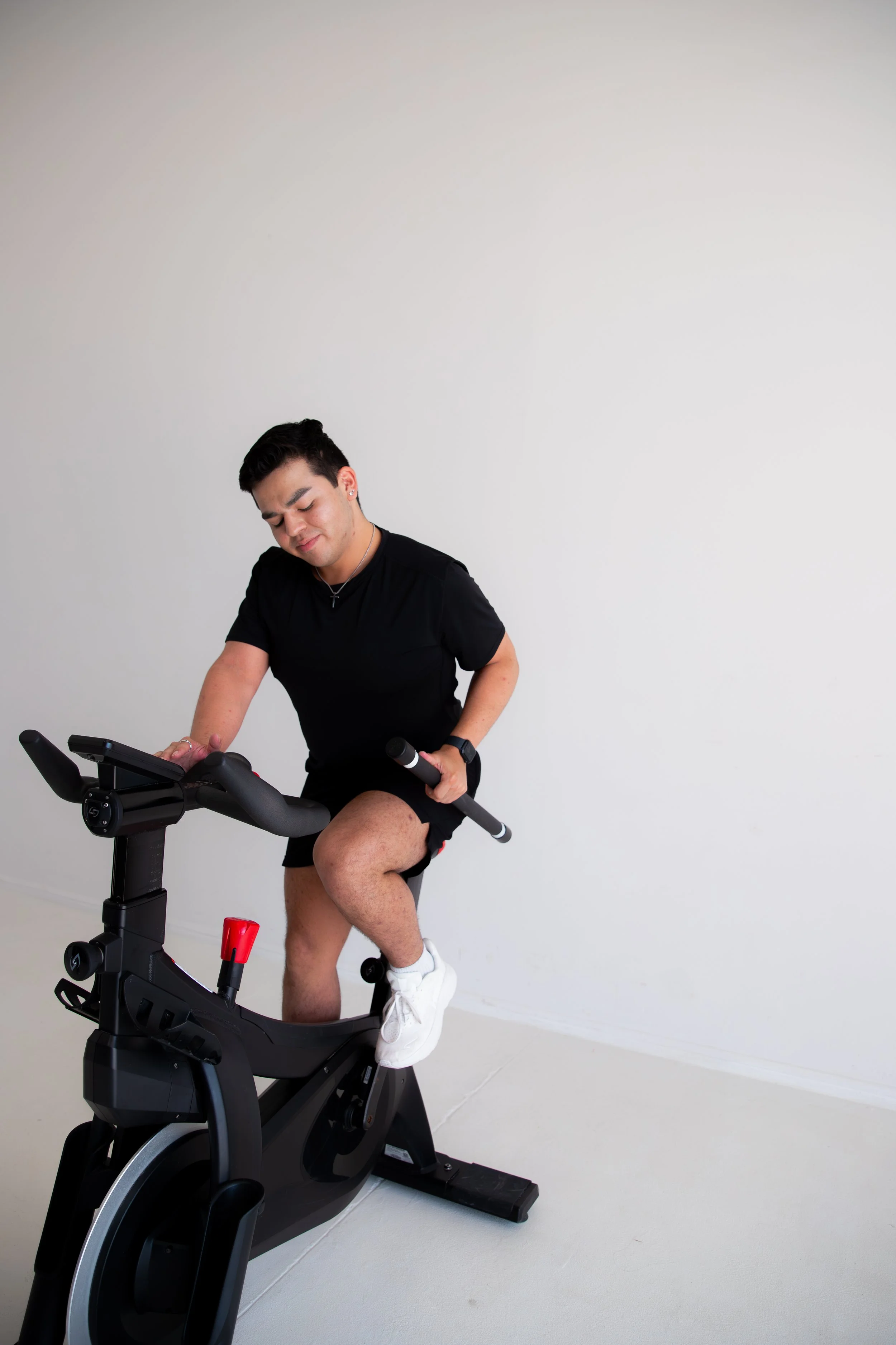 Young man wearing black t-shirt, black shorts, and white sneakers, working out on a stationary exercise bike indoors with a plain white wall background.
