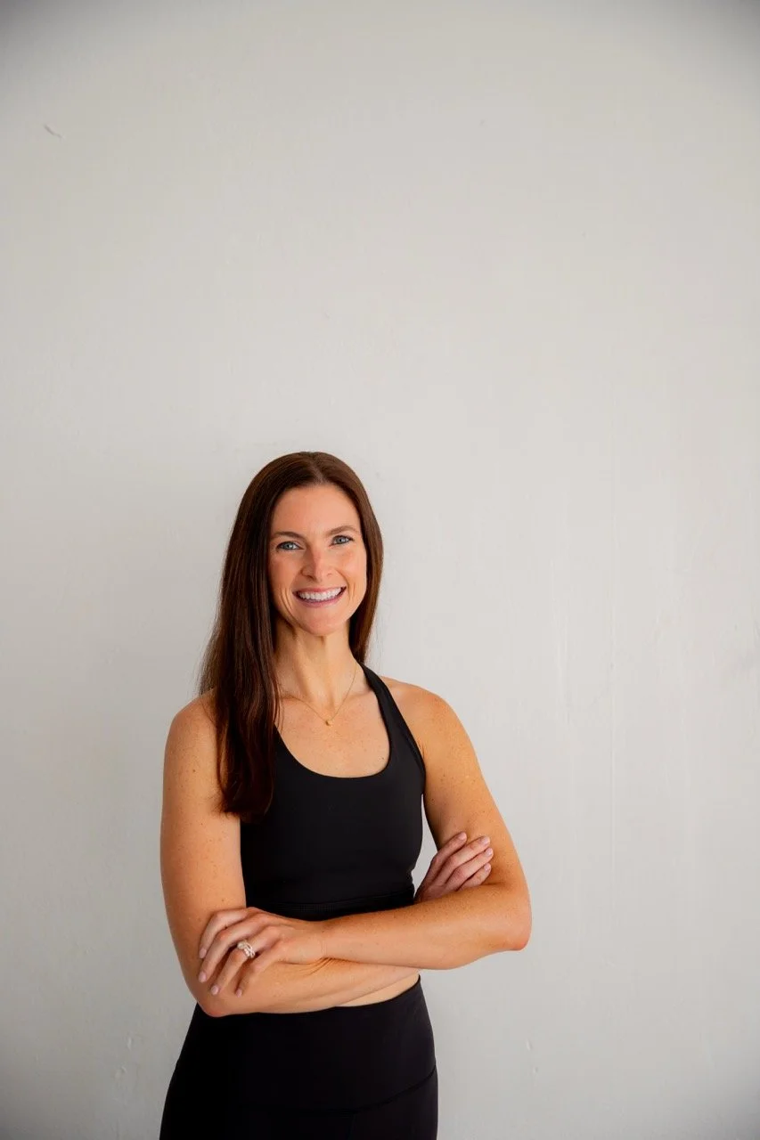 A smiling woman with long brown hair wearing a black workout top and black leggings, standing against a plain white wall with arms crossed.