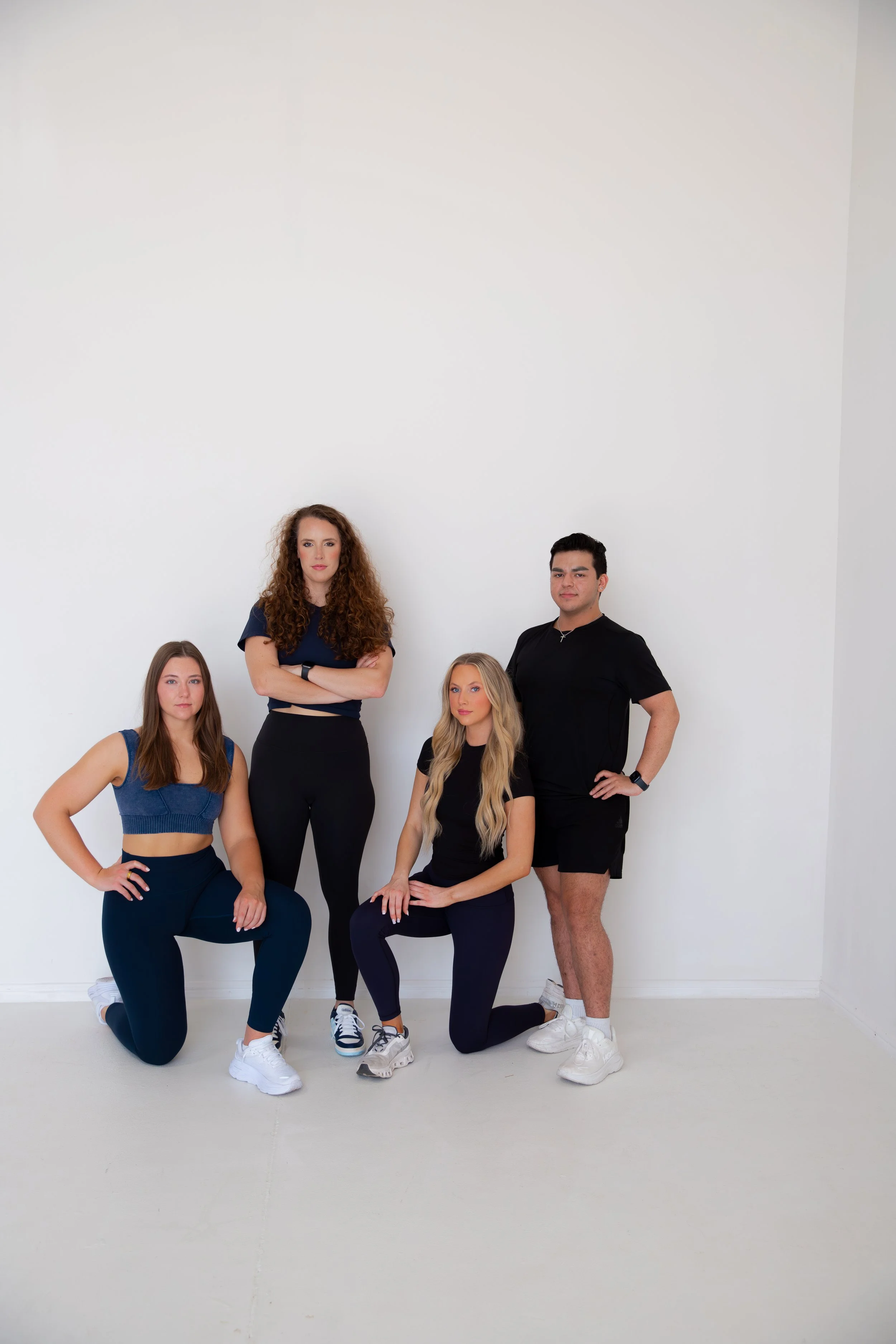 Four young adults in athletic wear posing against a white wall, with two kneeling and two standing.