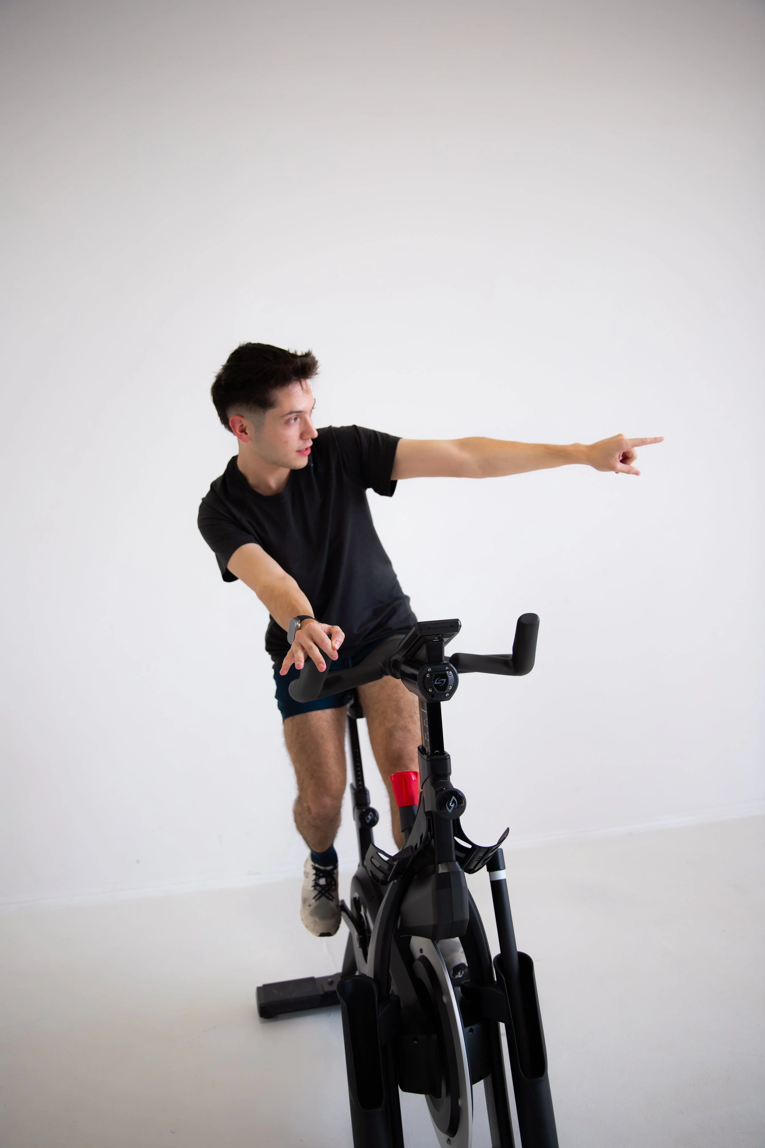 Young man in black t-shirt and shorts exercising on an exercise bike, pointing to the right.