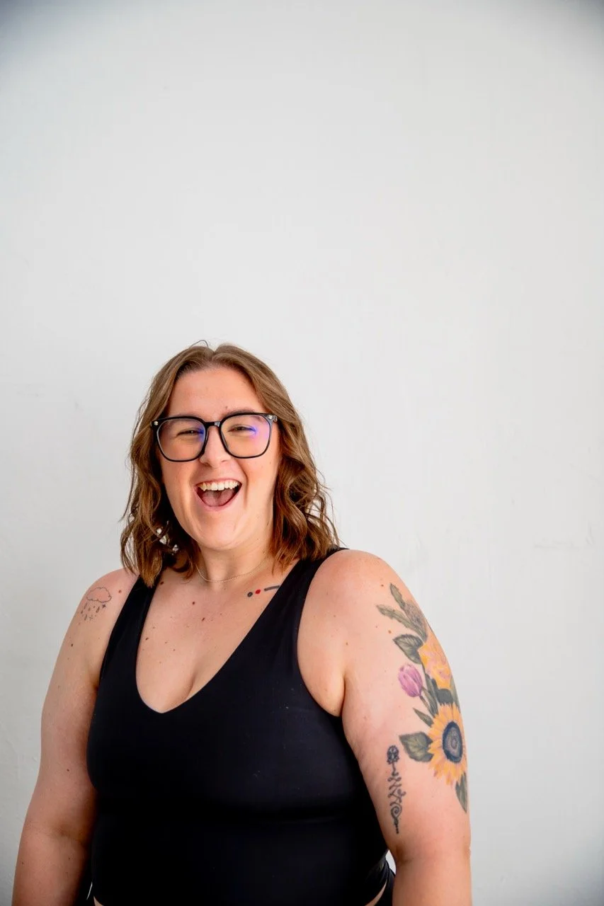 A woman with glasses, wavy brown hair, and multiple tattoos, smiling and laughing, wearing a black tank top against a plain white background.