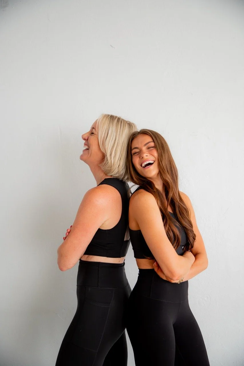 Two women in black athletic outfits standing back-to-back smiling and laughing against a white wall.