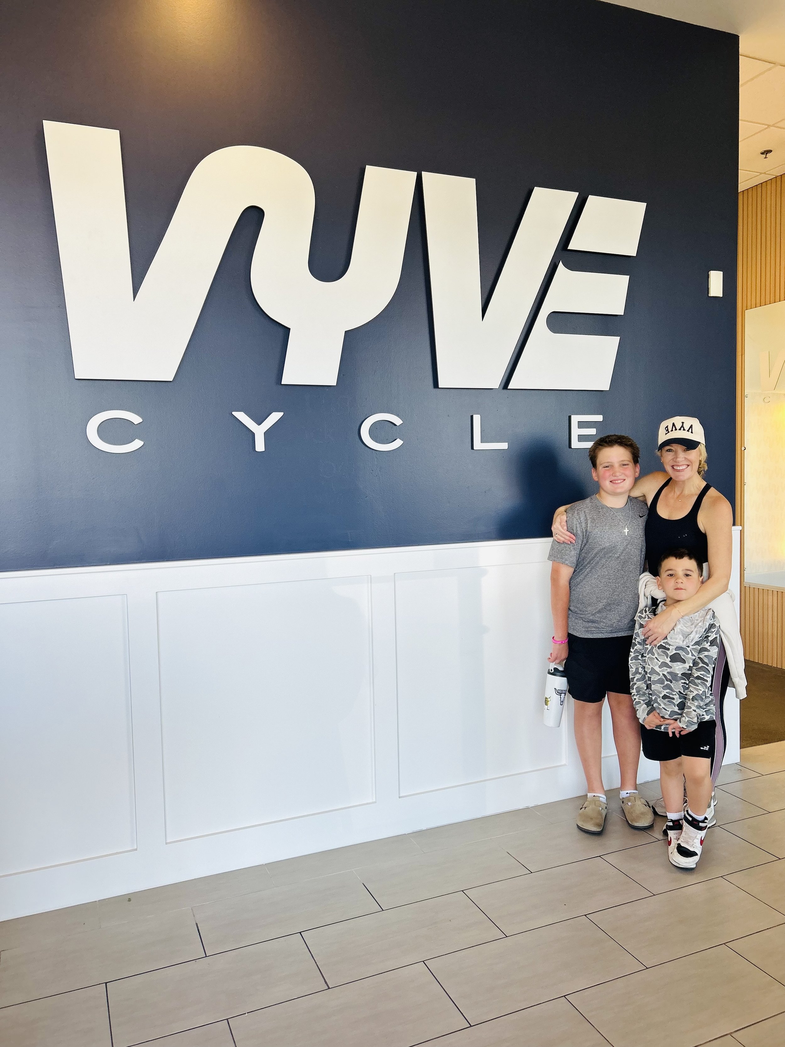 A woman, Michelle, owner of VYVE Cycle and two children standing in front of a navy blue wall with the words ' VYVE CYCLE' displayed in large white letters. T