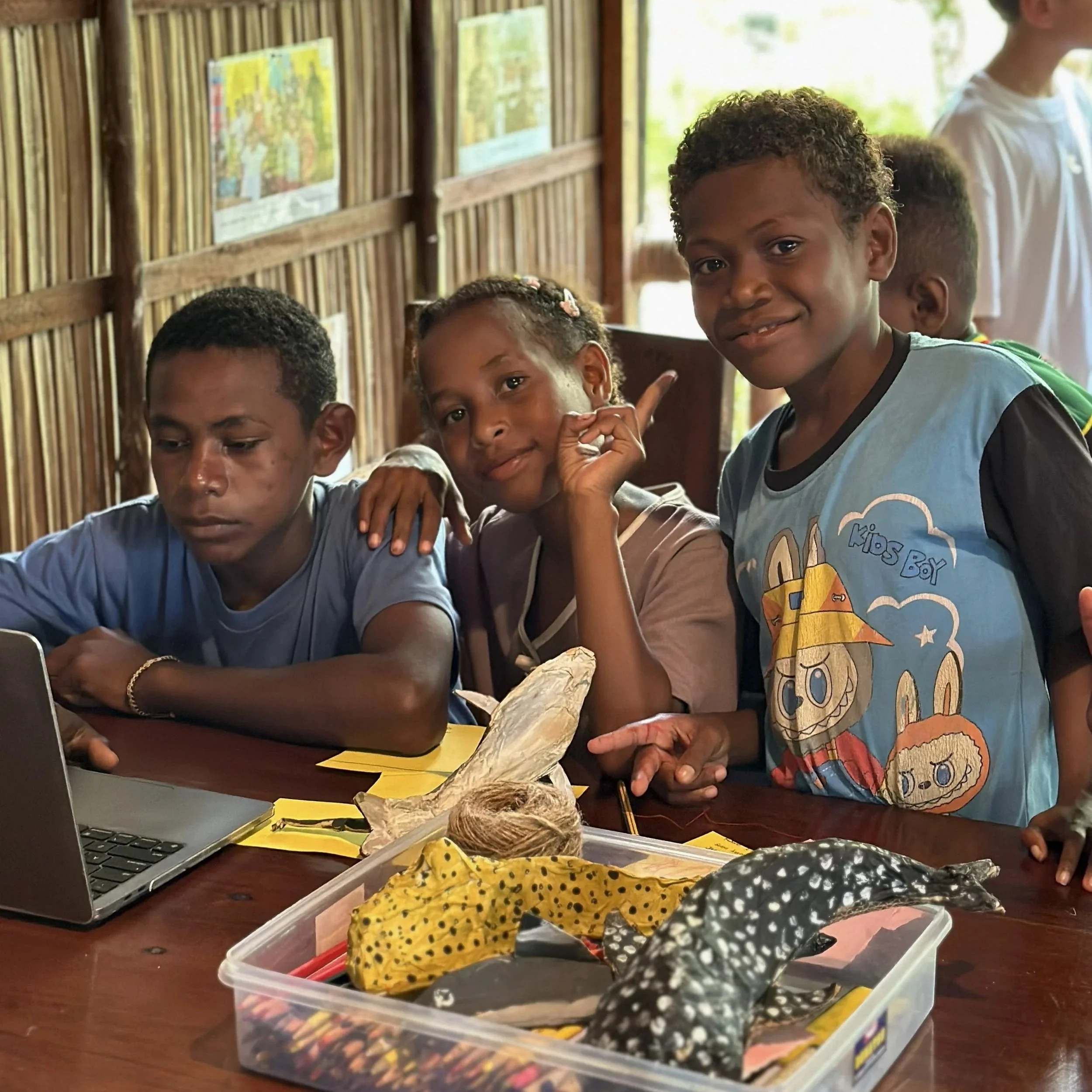 Three young students sitting at a wooden table at the Raja Ampat SEA Centre with a laptop and craft supplies, smiling and looking at the camera inside a wooden structure with colorful maps on the wall.