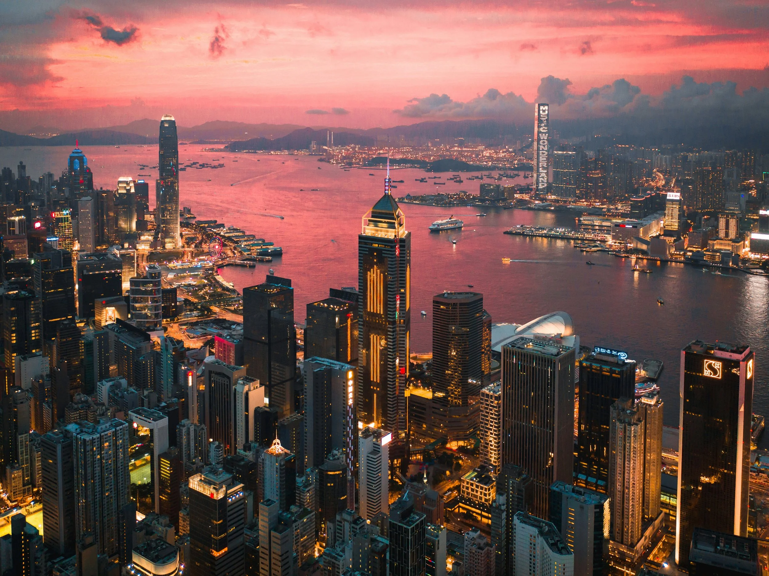 Aerial view of Hong Kong skyline at sunset with illuminated skyscrapers and Victoria Harbour.