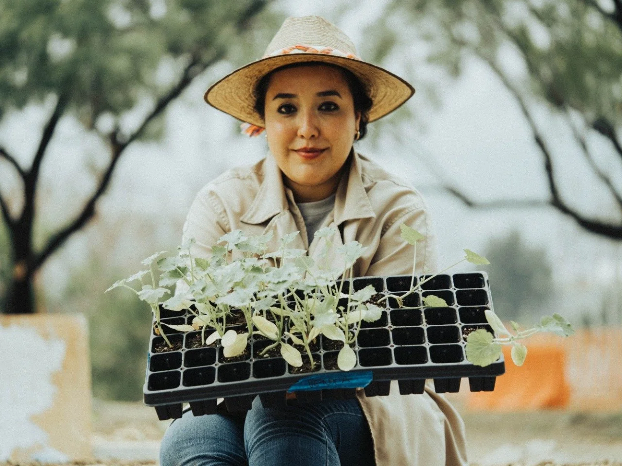 Mujer joven con sombrero de paja sosteniendo una bandeja con plántulas en un fondo de árboles y cielo.