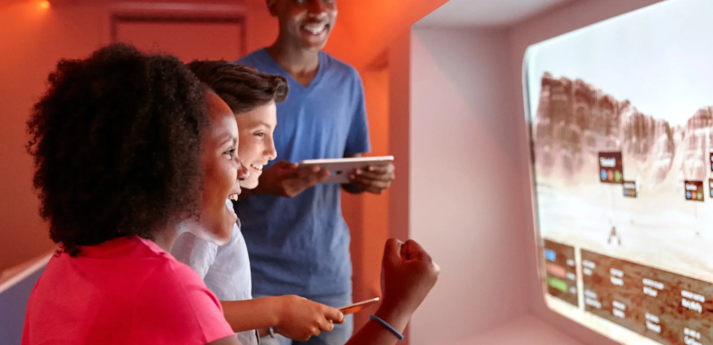 Three children, two girls and one boy, are looking excited at a large screen showing a video game in a room with orange and white walls. An adult man stands behind them smiling.