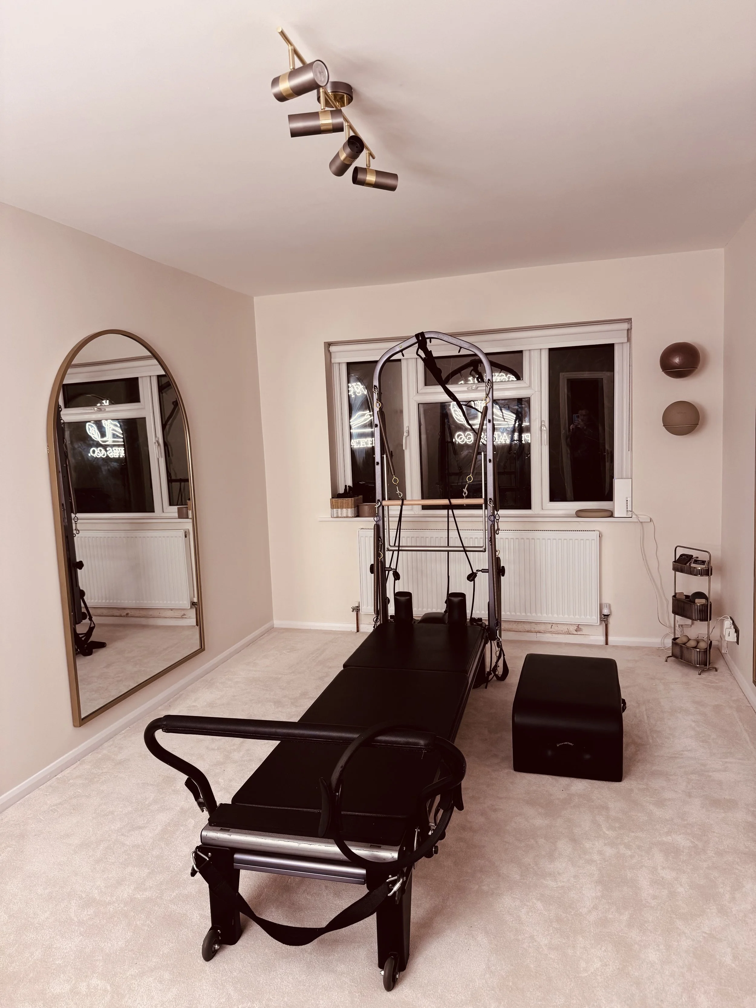 A home exercise room with a Pilates reformer, a large mirror on the wall, a window, and a storage rack with rolled towels.