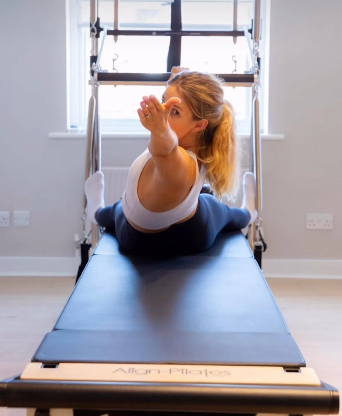 A woman doing a Pilates exercise on a reformer machine, lying on her stomach and stretching her right arm forward.