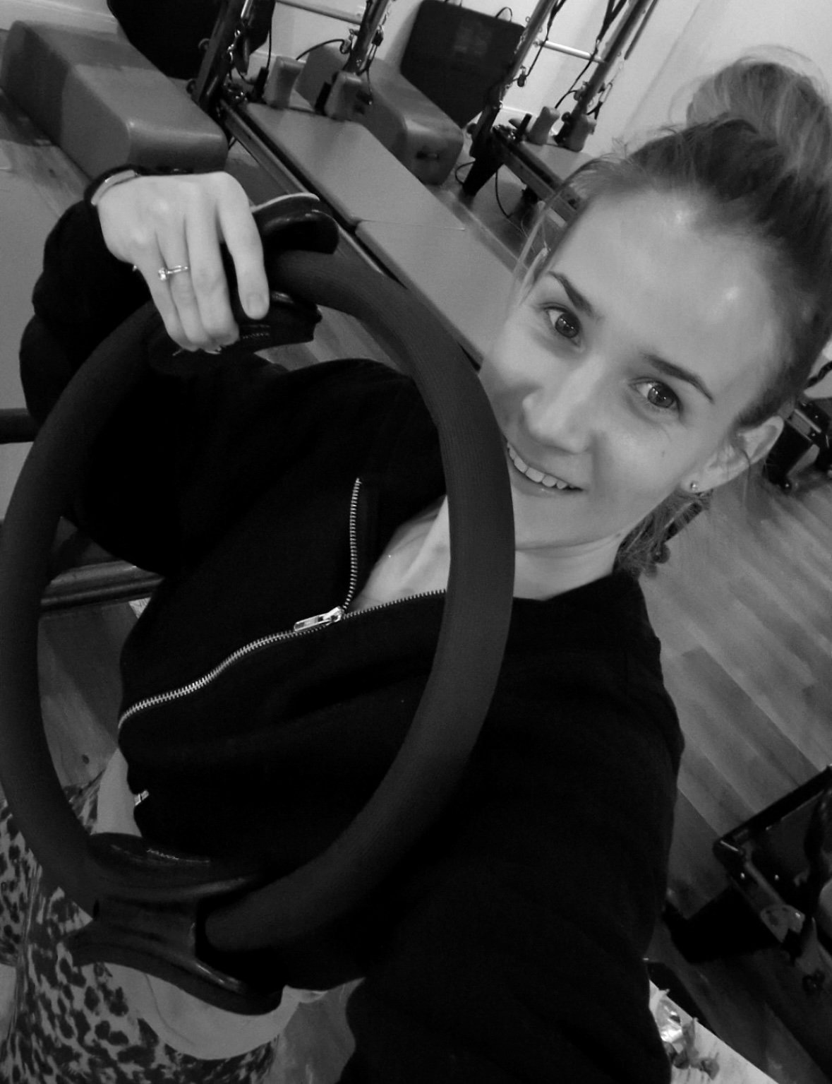 Black and white photo of a smiling woman holding a bicycle handlebar, with gym equipment in the background.