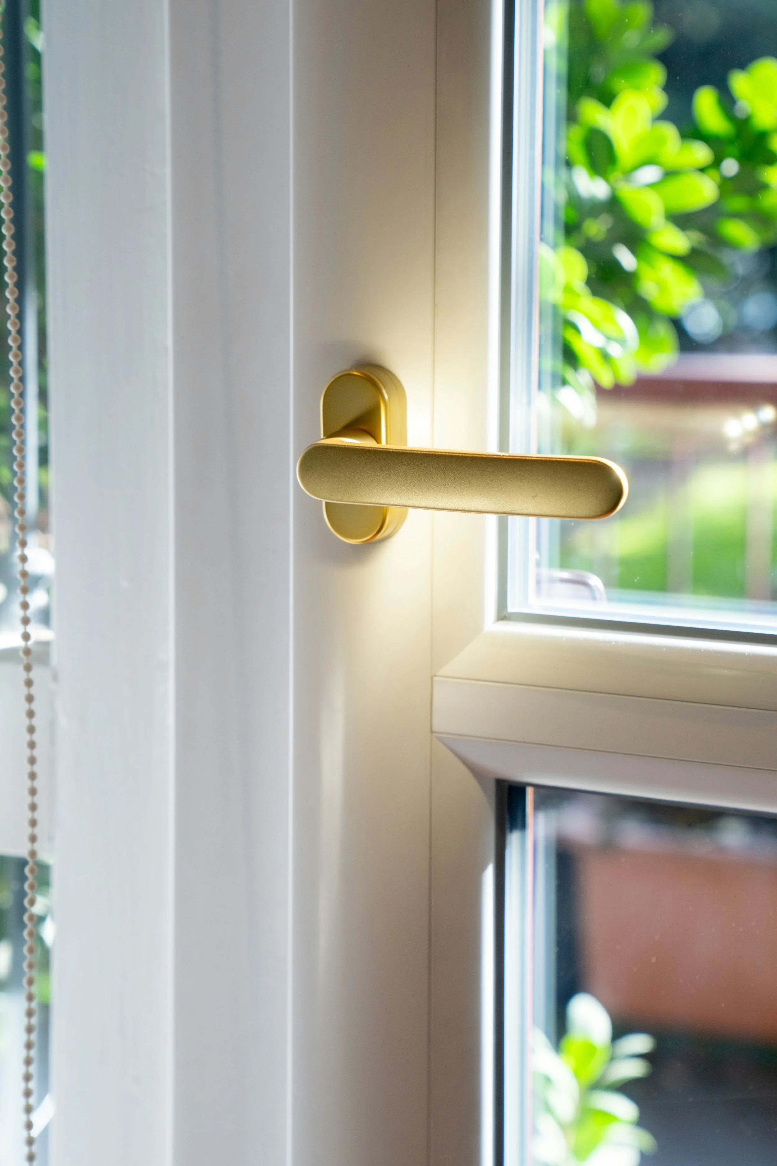 Close-up of a gold-colored door handle on a white door, with sunlight coming through a window showing green foliage outside.