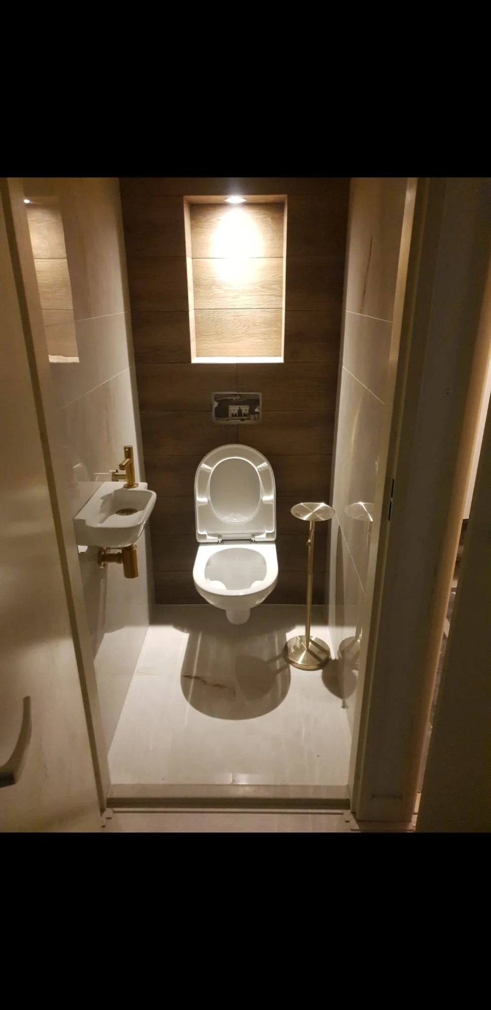 Small modern toilet room with wood paneling on the back wall, a white toilet with a partially open lid, a small wall-mounted sink with a gold faucet, a gold toilet paper stand, and recessed lighting.