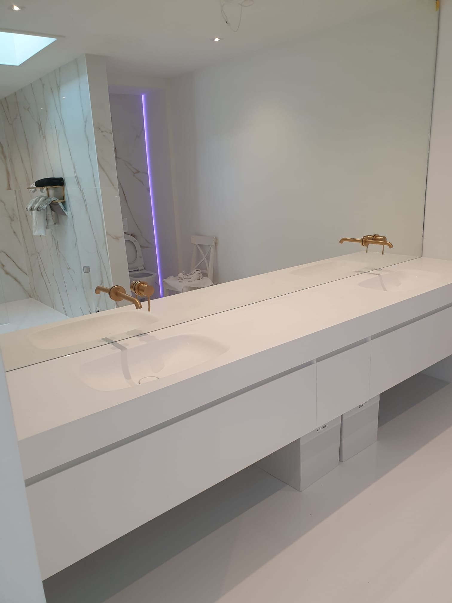 White double vanity with two sinks and gold fixtures, large mirror above, in a modern minimalistic bathroom with marble accents and purple LED lighting in the background.