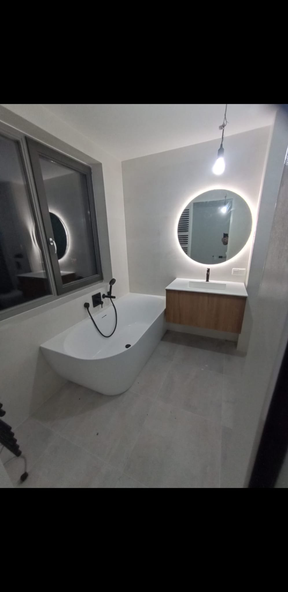 Modern bathroom with a freestanding bathtub, a large round backlit mirror, a wooden vanity with a white sink, and a window with a round mirror reflection.