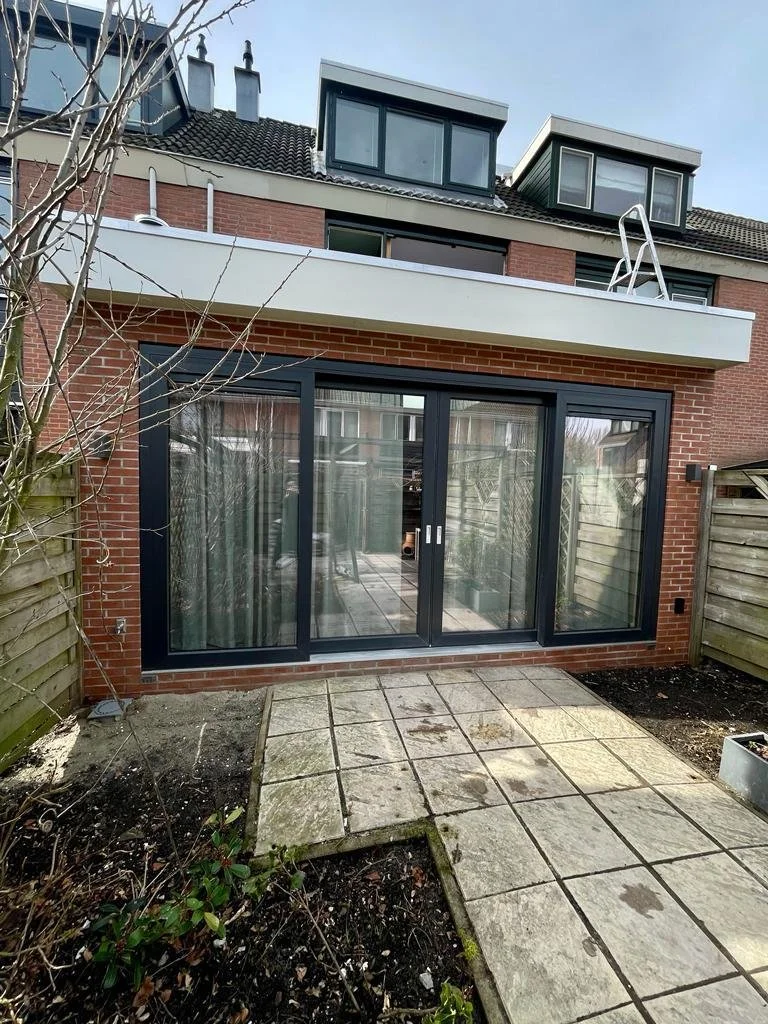 Backyard patio with stone tiles leading to large glass sliding doors, brick exterior house, and garden fence. Upper balcony with a ladder visible on the roof.