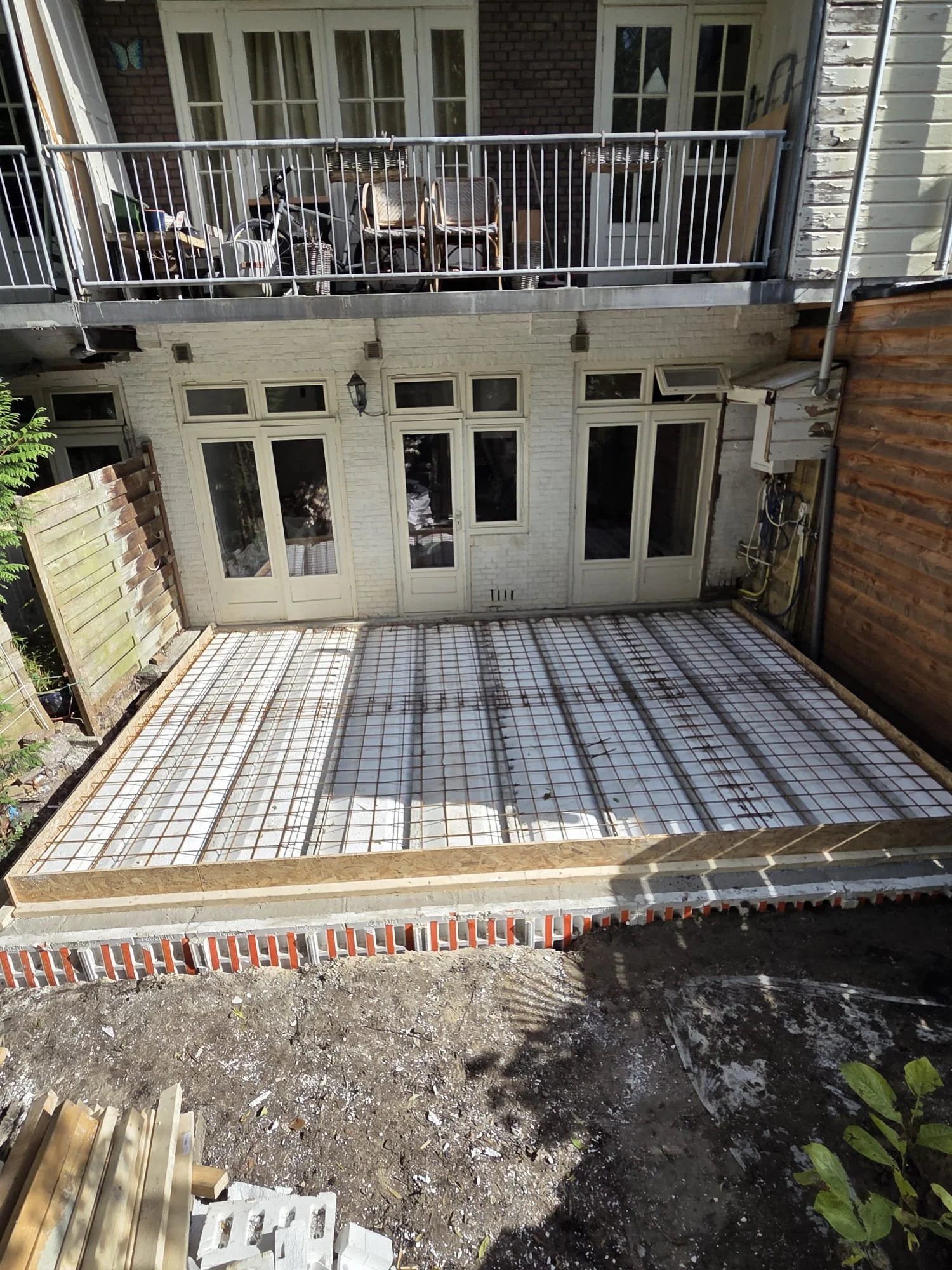 Construction site of a backyard patio with rebar grid, in front of the house, which has white door and windows, and an upper balcony with chairs and outdoor furniture.