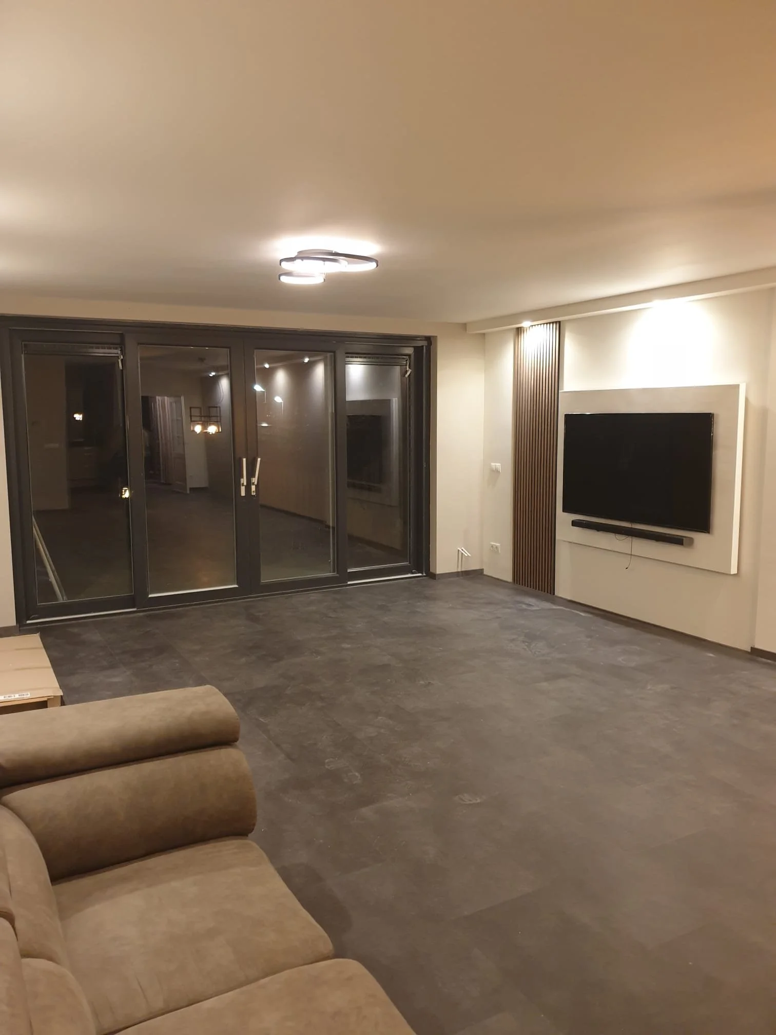 A modern living room with a beige sofa in the foreground, a large flat-screen TV mounted on the wall to the right, and floor-to-ceiling glass sliding doors. The room has dark flooring and minimal decor.