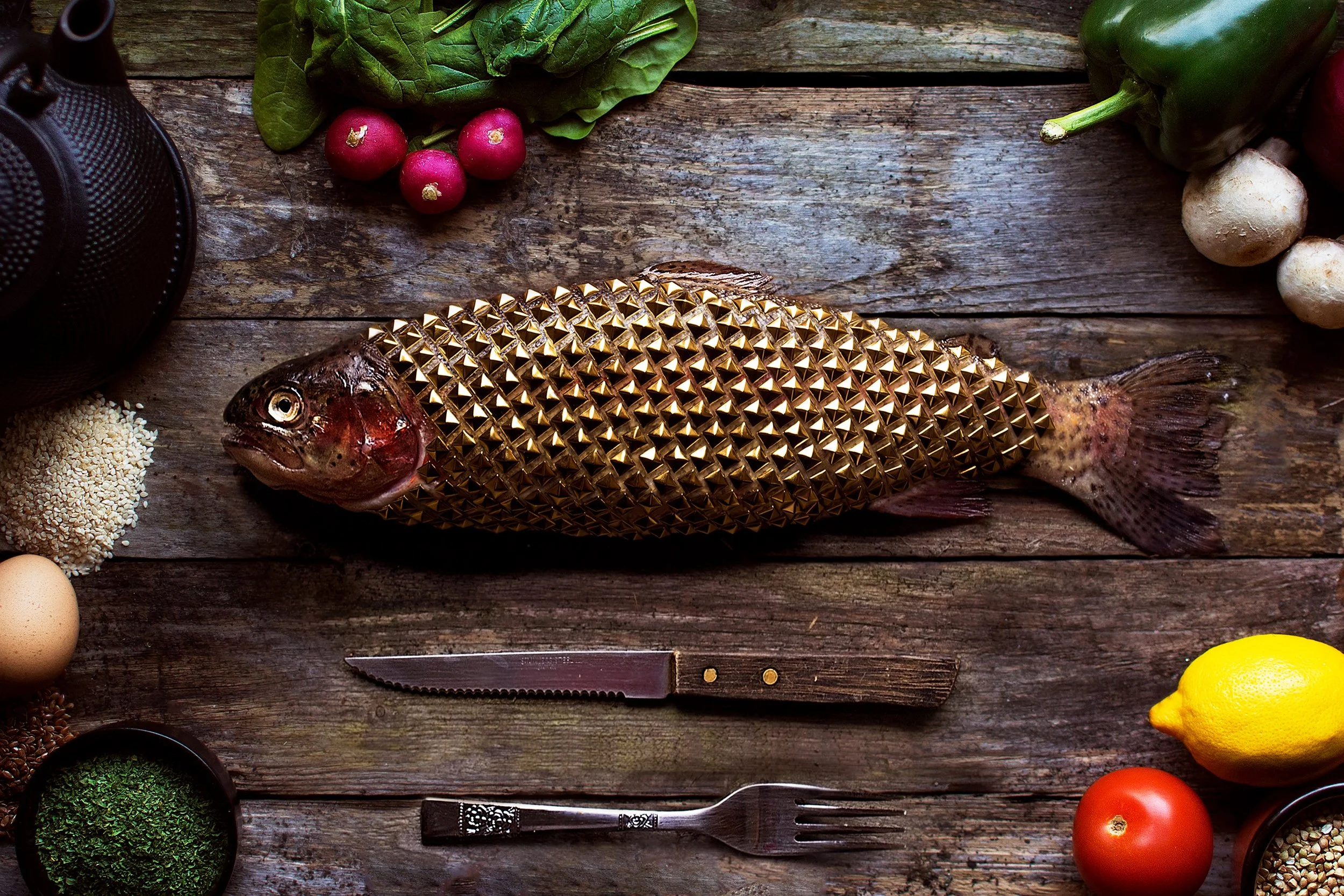 A fish with a golden spiked exterior lying on a rustic wooden table, surrounded by vegetables, eggs, herbs, a lemon, a tomato, and kitchen utensils.