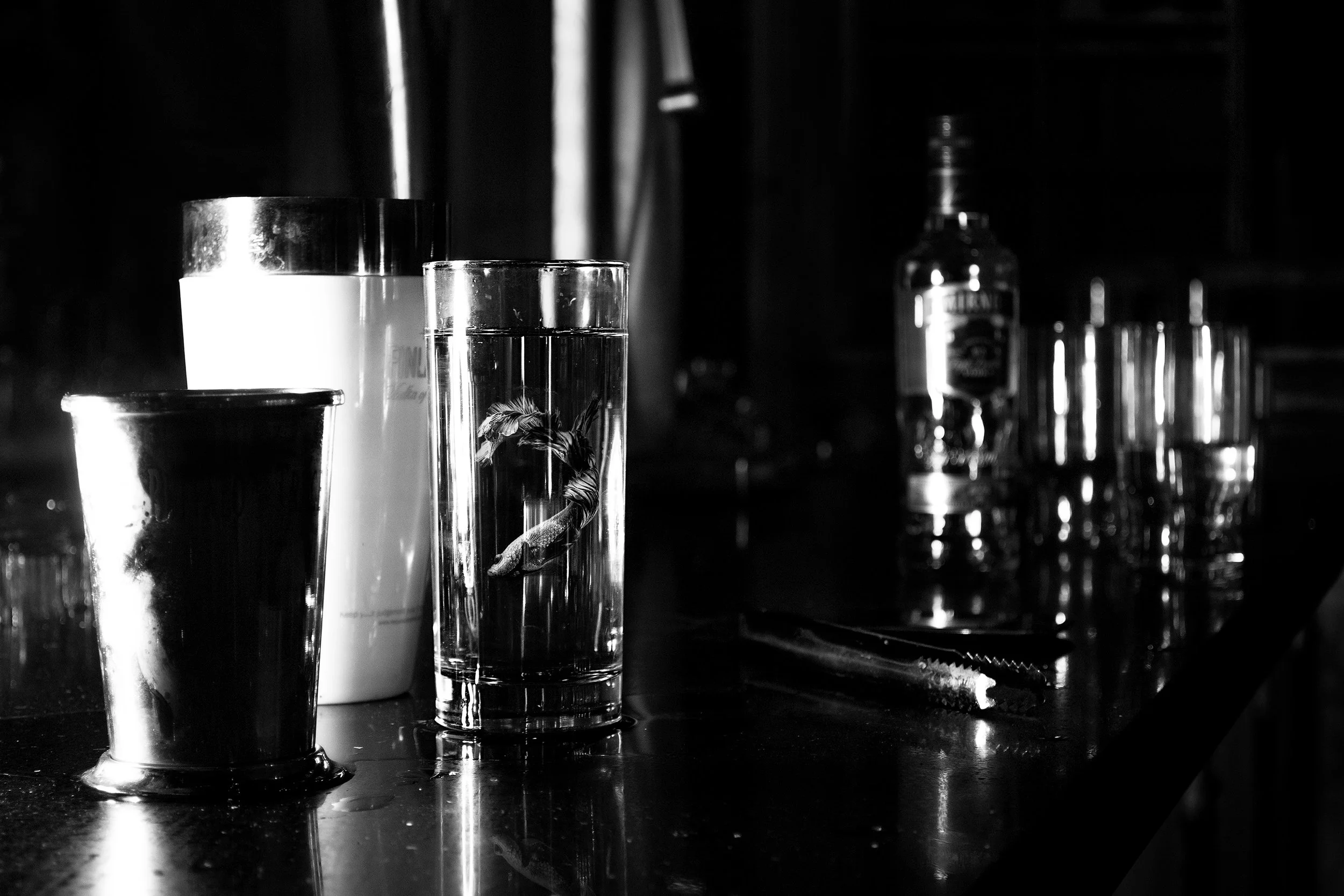 An array of drink glasses and bottles on a bar or counter, including a glass with a shrimp, a shot glass, a pint glass, a bottle of alcohol, and a cocktail shaker, in black and white.
