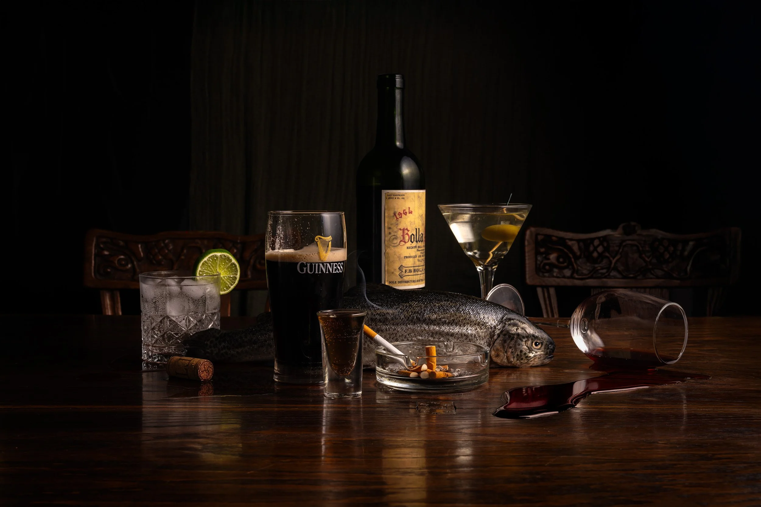 A dark tabletop featuring a fish, cigarette pack, glass of red wine spilled on the table, pint of Guinness, and bottles of alcohol, with various drinks and garnishes in the background.