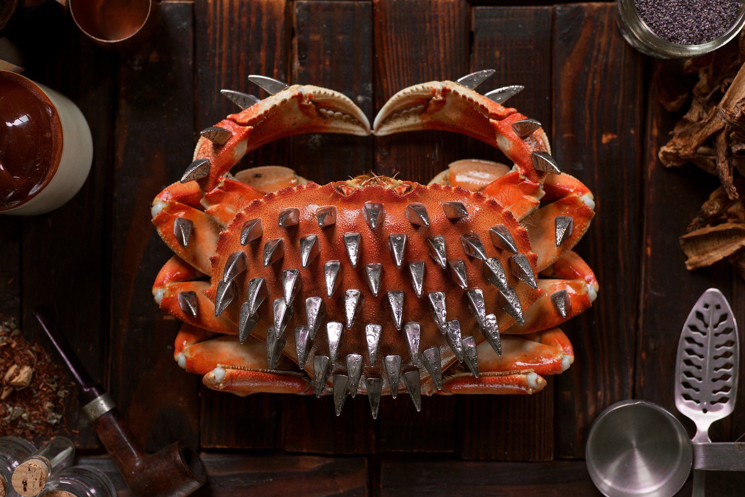 A spiny crab with orange shell and large silver claws placed on a dark wooden surface surrounded by cooking tools and ingredients.