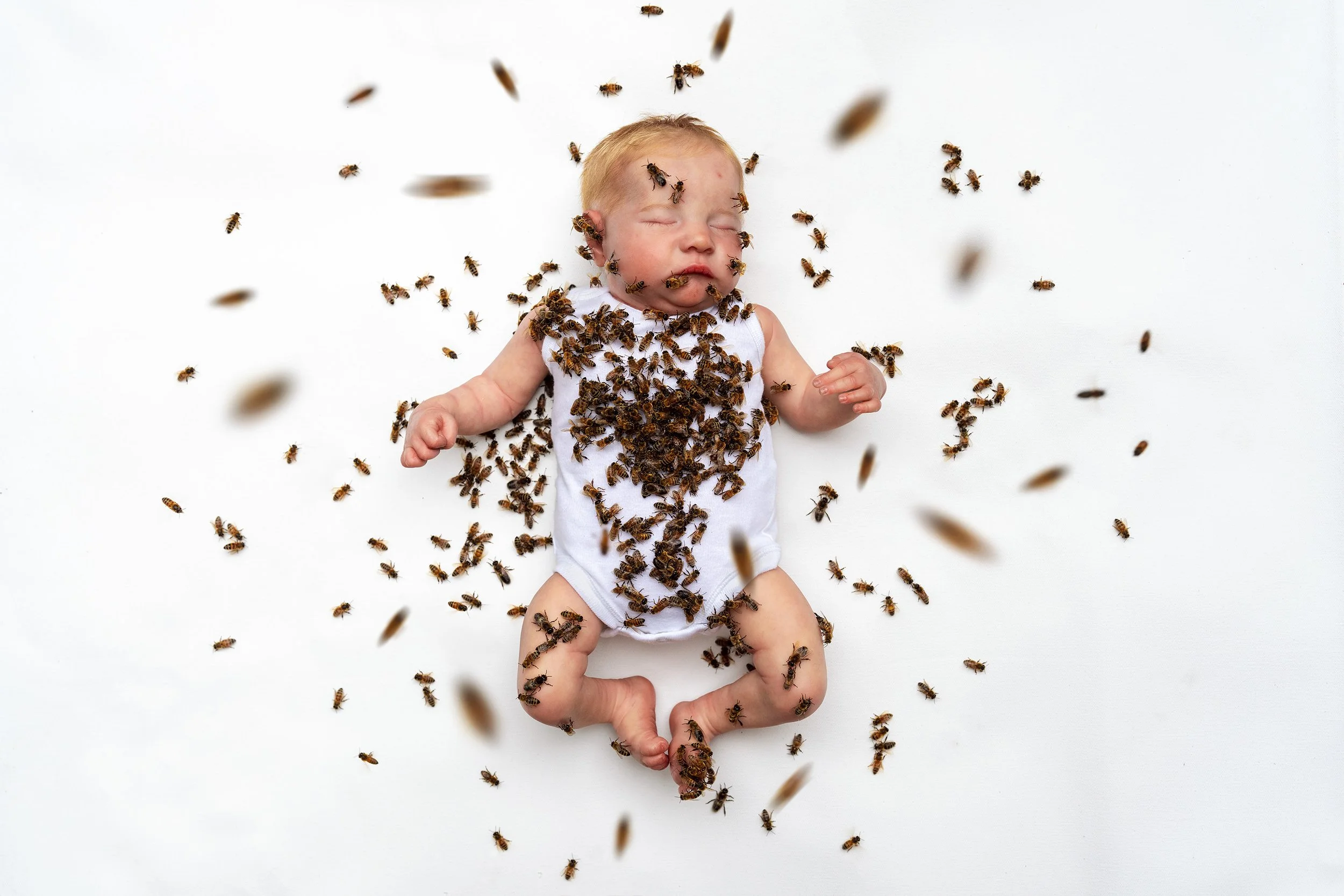 Baby lying on a white surface with bees all over him and around him, sleeping peacefully.