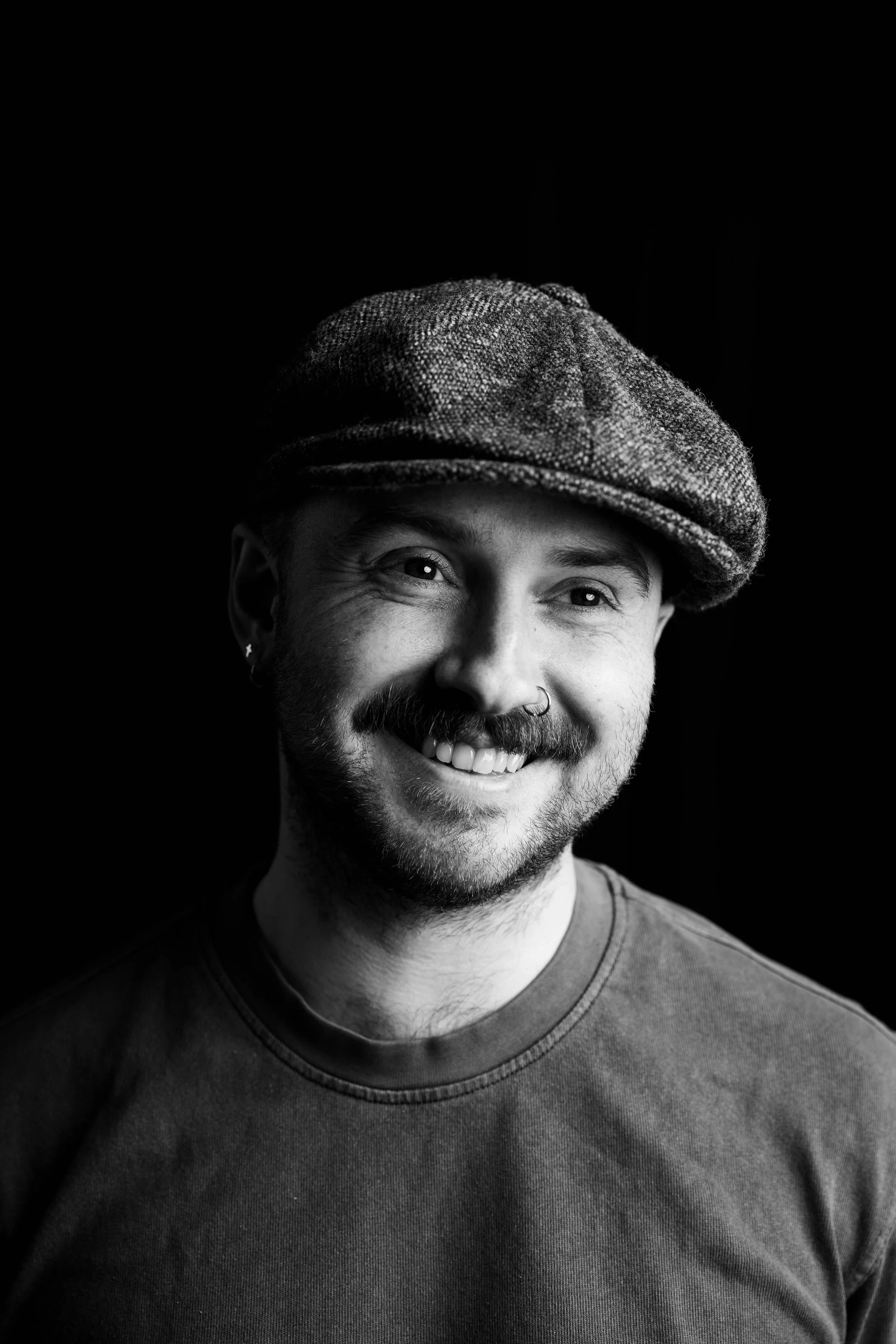 Black and white portrait of a smiling man with a beard, mustache, and earring wearing a flat cap and a T-shirt, against a dark background.