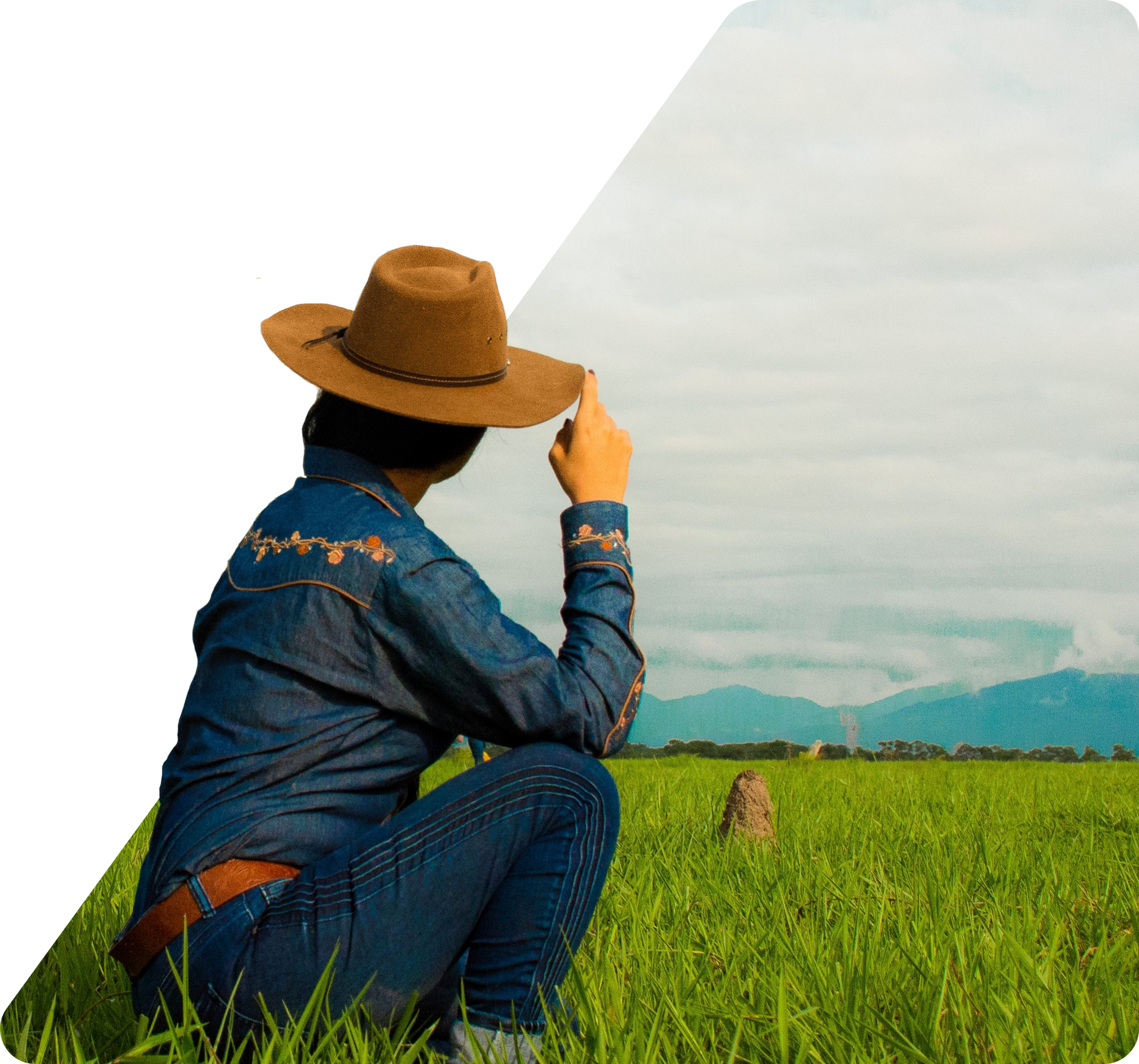 Woman wearing a wide-brimmed hat and denim jacket crouching in a grassy field with mountains and a cloudy sky in the background.