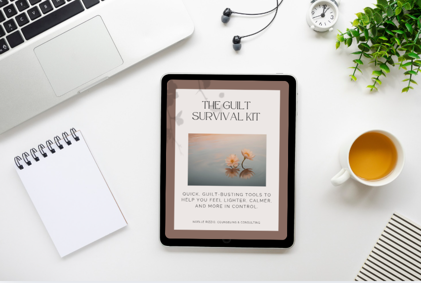 Tablet displaying a book cover titled 'The Working Moms Guilt Survival Kit' with an image of two pink water lilies on water, surrounded by a white desk, a cup of tea, a notebook, a plant, and electronic accessories.