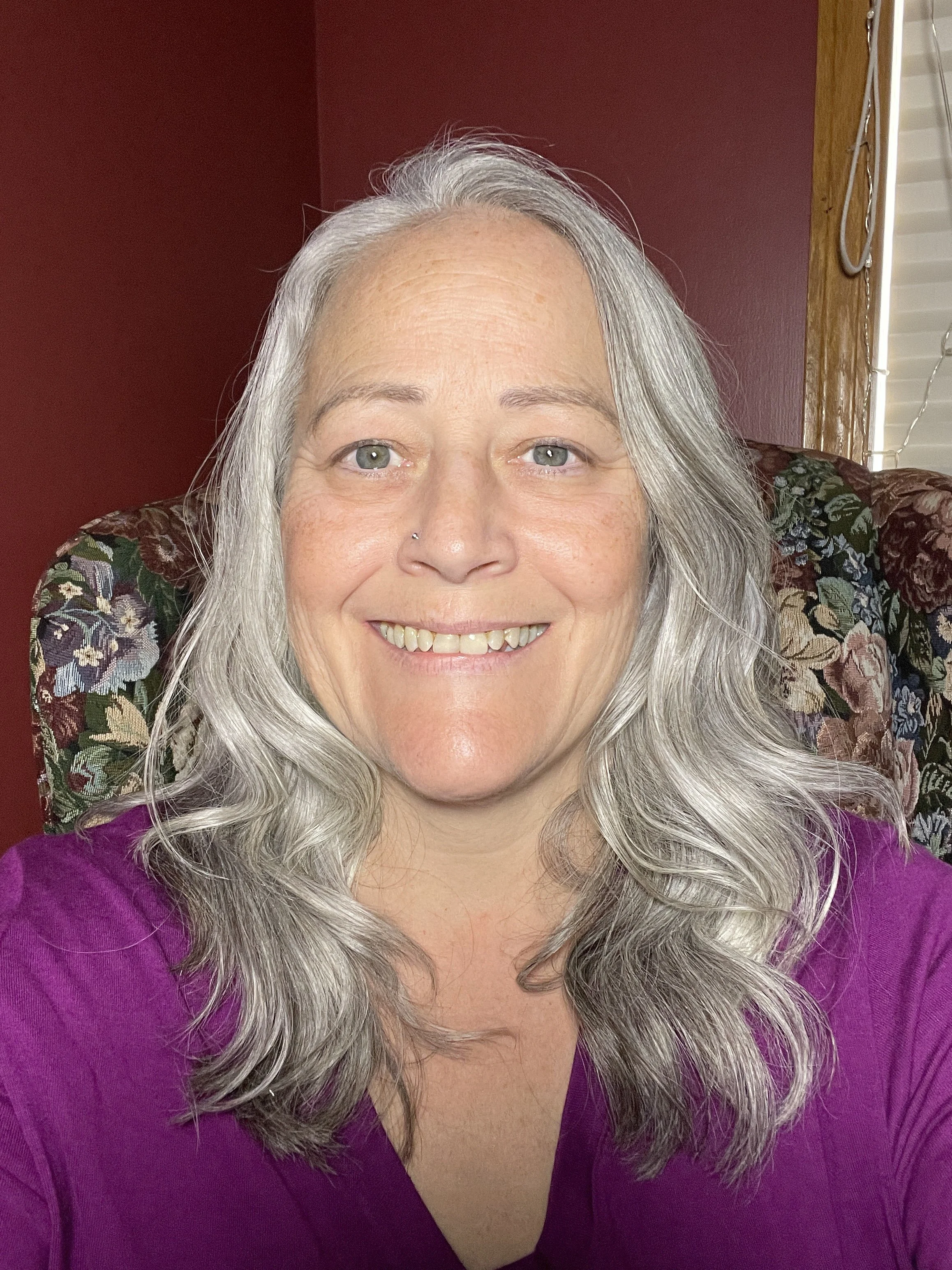 A smiling woman with long gray hair, light skin, and a purple top, seated on a floral-patterned chair indoors.