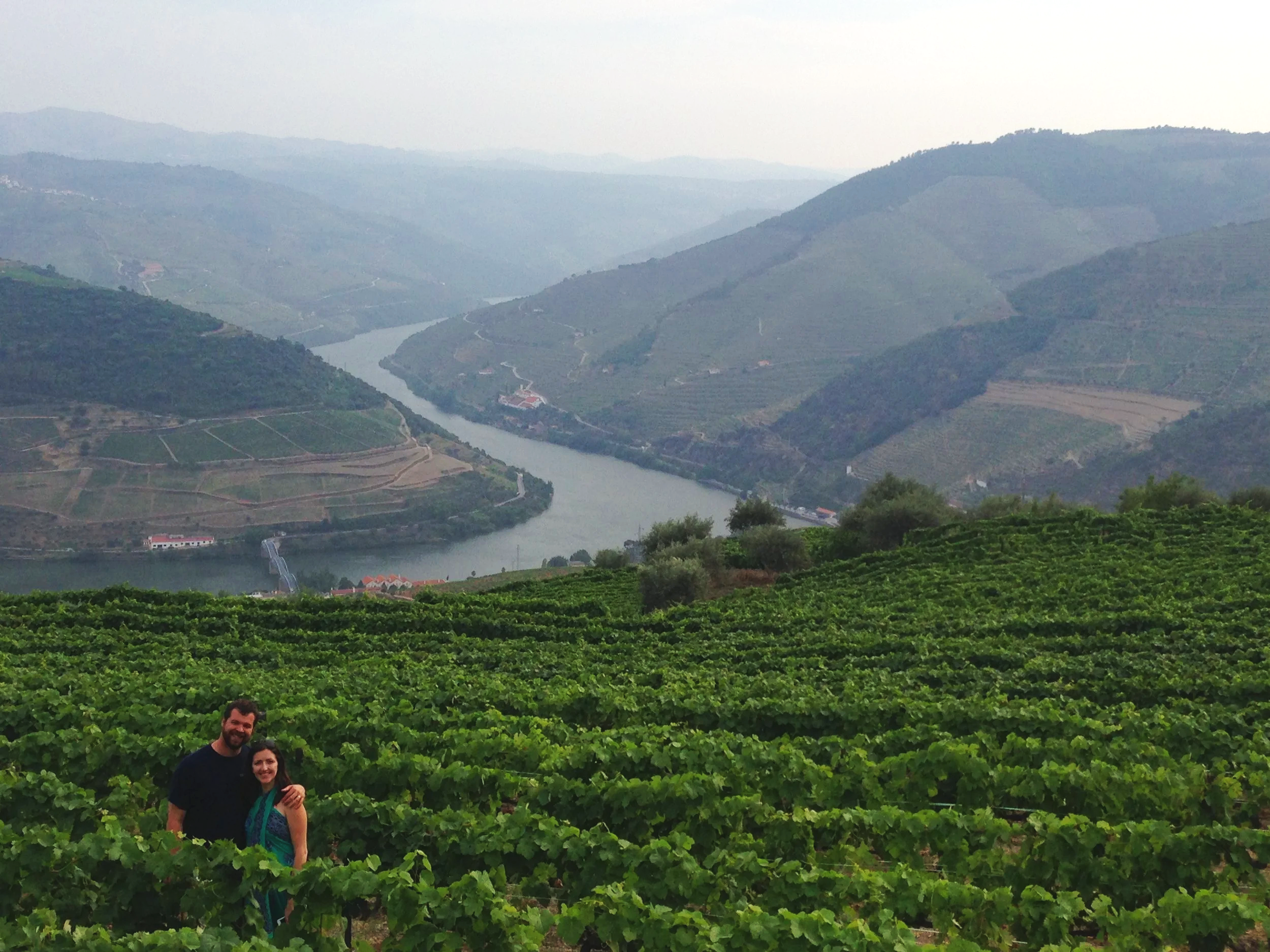 A couple standing in lush green vineyard with rolling hills and a river in the background, under a hazy sky.
