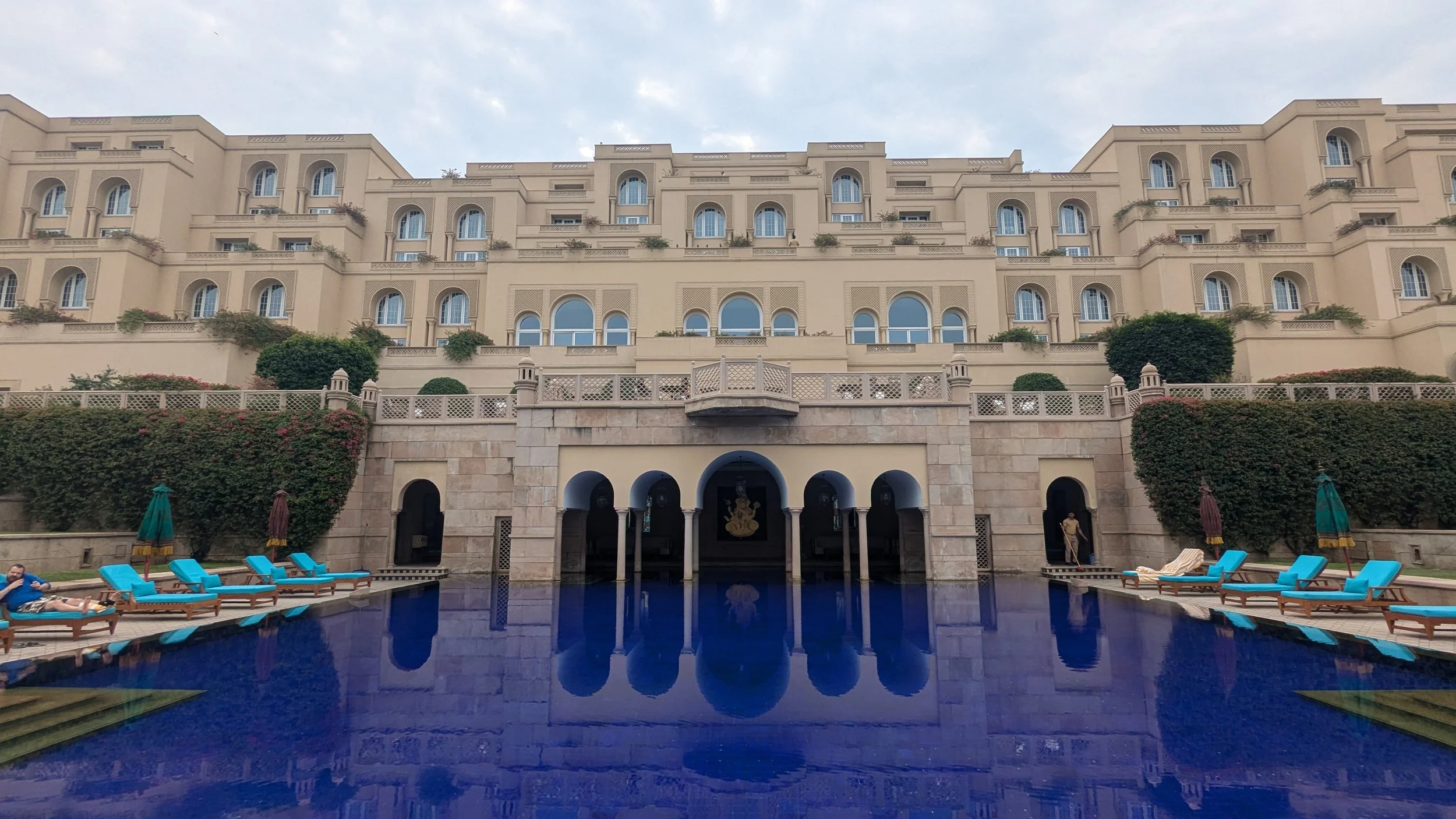 A large, elegant hotel with an ornate beige facade and many arched windows, overlooking a deep blue swimming pool surrounded by lounge chairs and umbrellas, with some greenery and a face statue in the background.