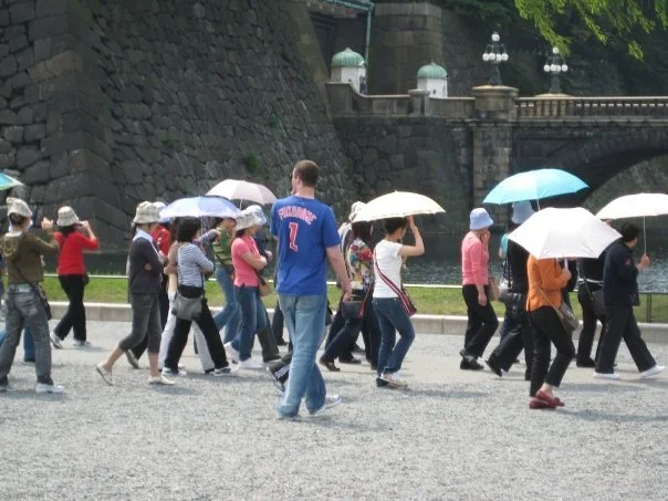 Group of people walking with umbrellas on a city street near a stone wall and a bridge.
