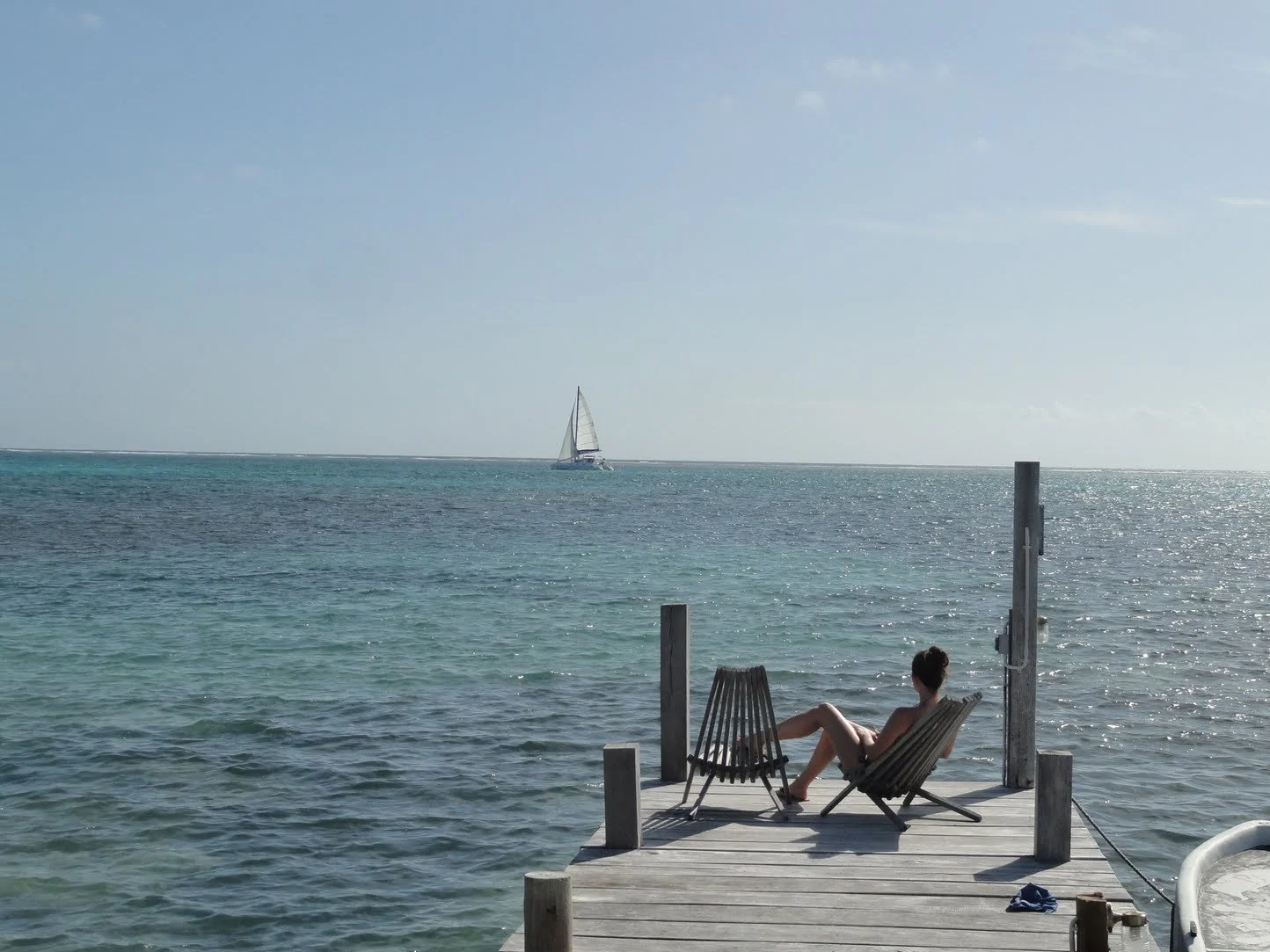 A woman relaxing on a wooden dock by the water, facing the ocean. A sailboat is visible in the distance on a sunny day with clear skies.