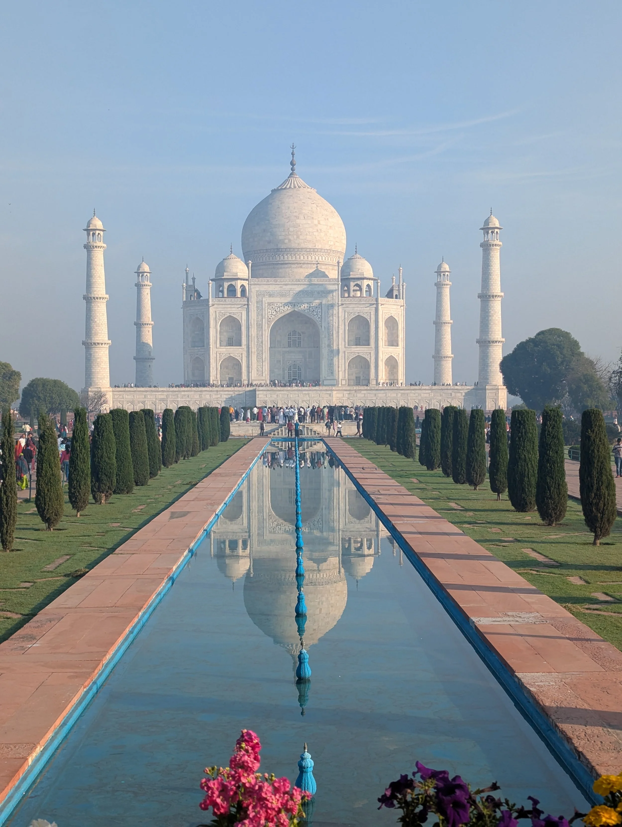 The Taj Mahal, a white marble mausoleum in India, is seen in this photo with clear blue sky. The view shows a reflecting pool leading to the monument, flanked by manicured bushes and colorful flowers in the foreground, with visitors near the entrance.