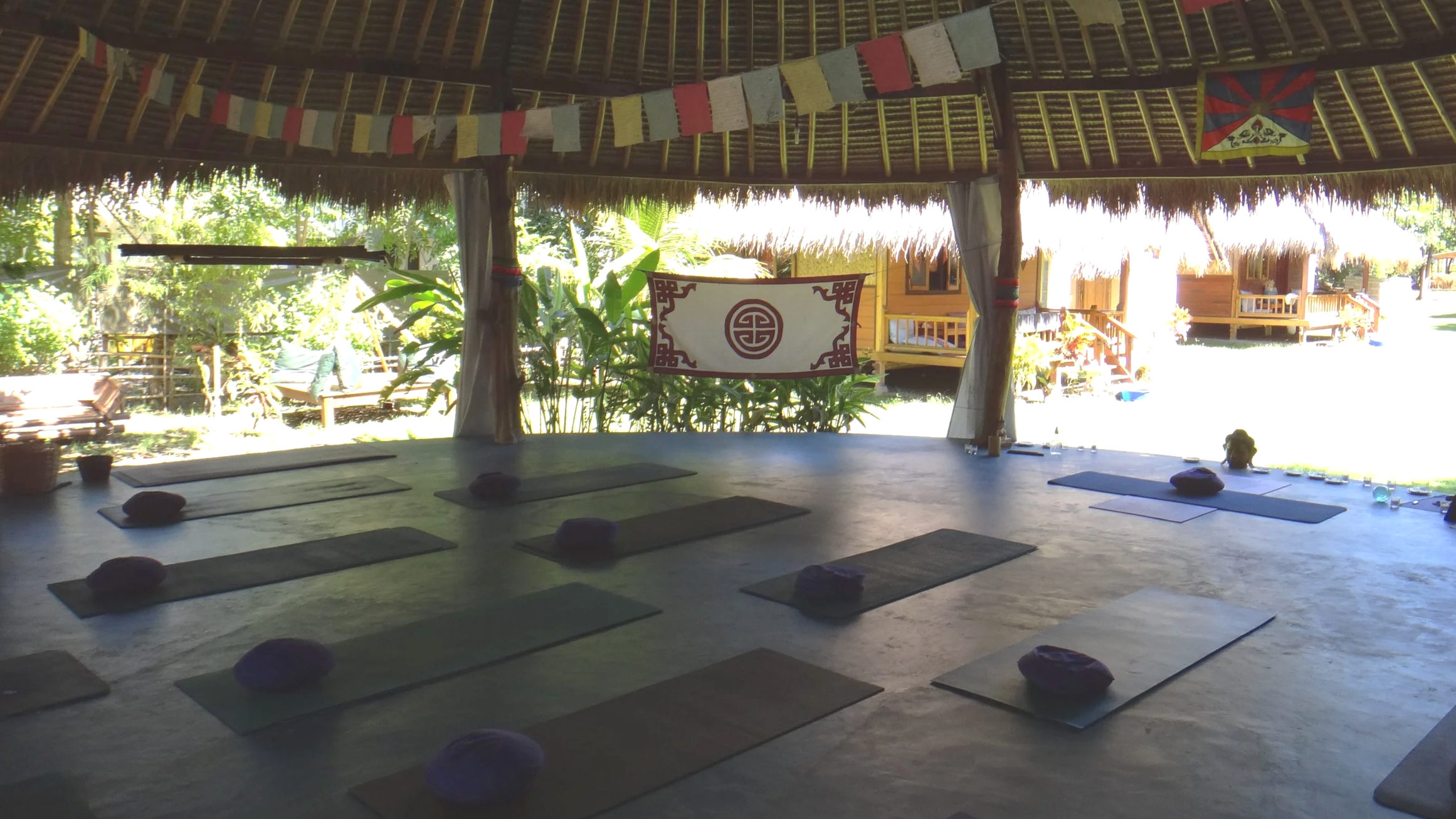 Interior of a meditation or yoga pavilion with 10 yoga mats placed on the floor, each topped with a meditation cushion. The pavilion has an open wall view of lush tropical greenery and traditional wooden structures in the background. Hanging decorations include colorful prayer flags and a banner with a symbol.