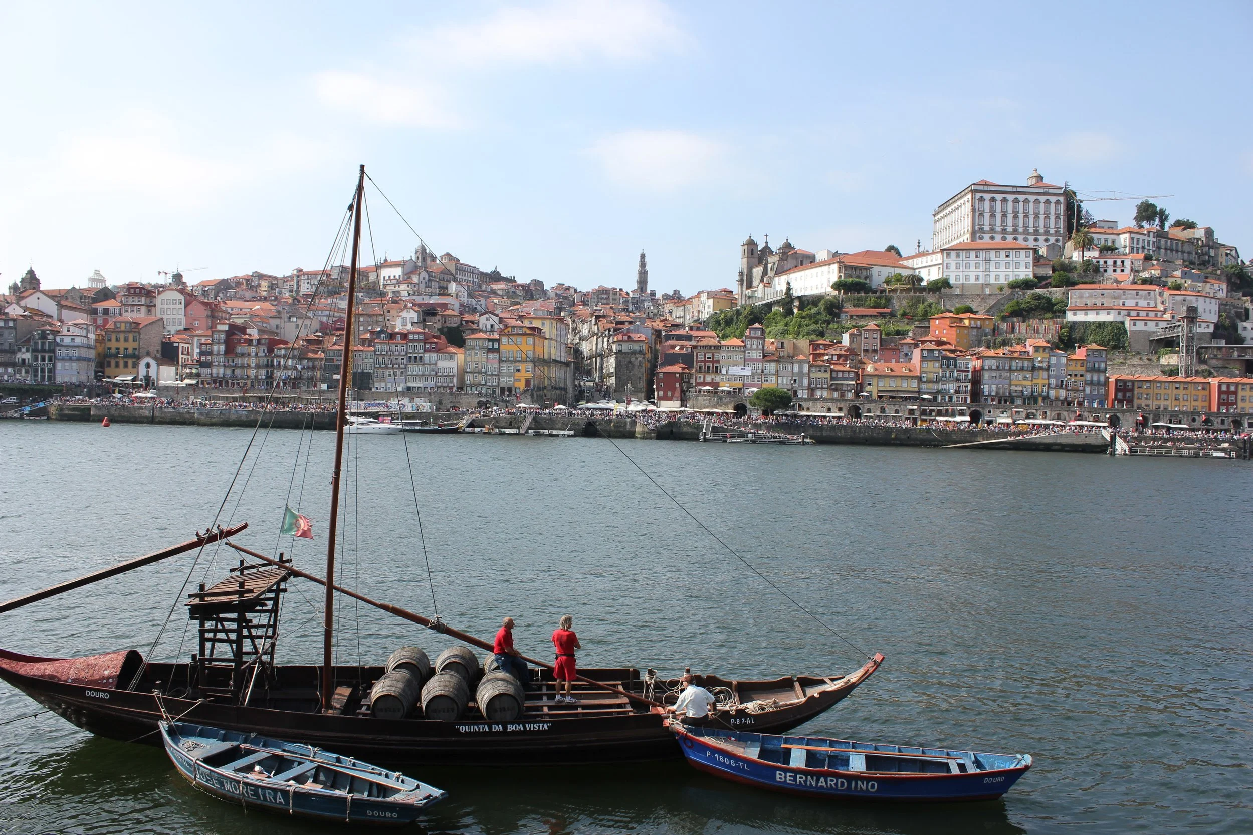 A traditional Portuguese boat floating on the river with barrels on board, and a scenic hillside city in the background with colorful buildings and historic landmarks.
