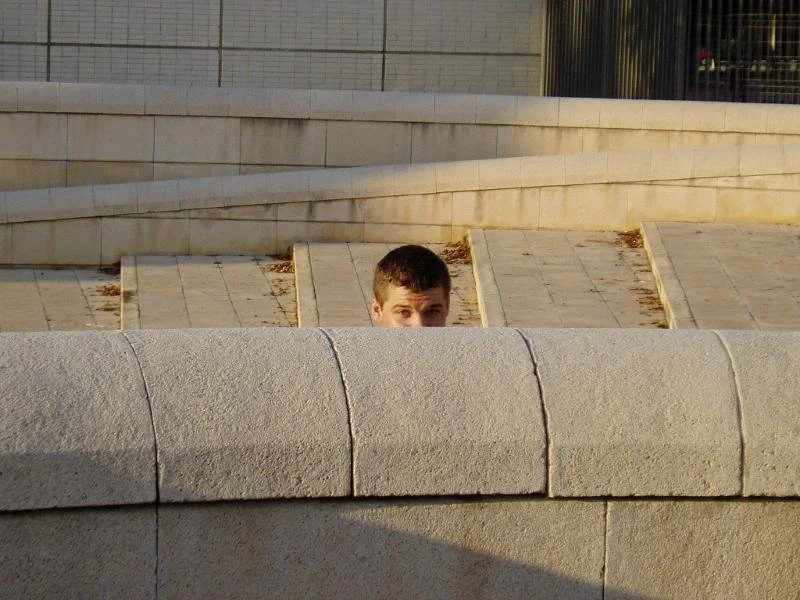 A young man peeking over a low stone wall on a pathway.