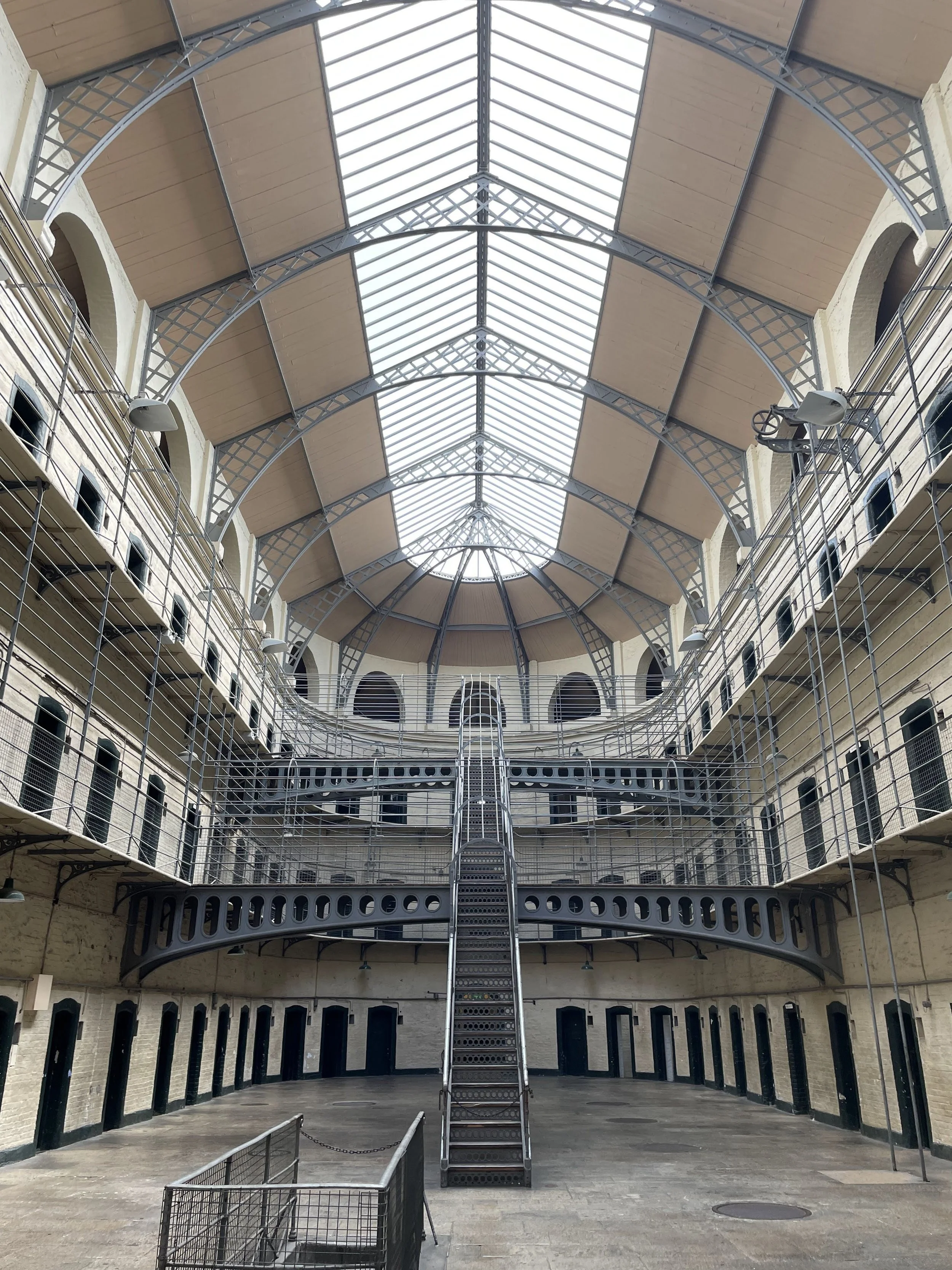 Interior of a historic prison with multiple levels of cells, metal walkways, and a large glass skylight ceiling.