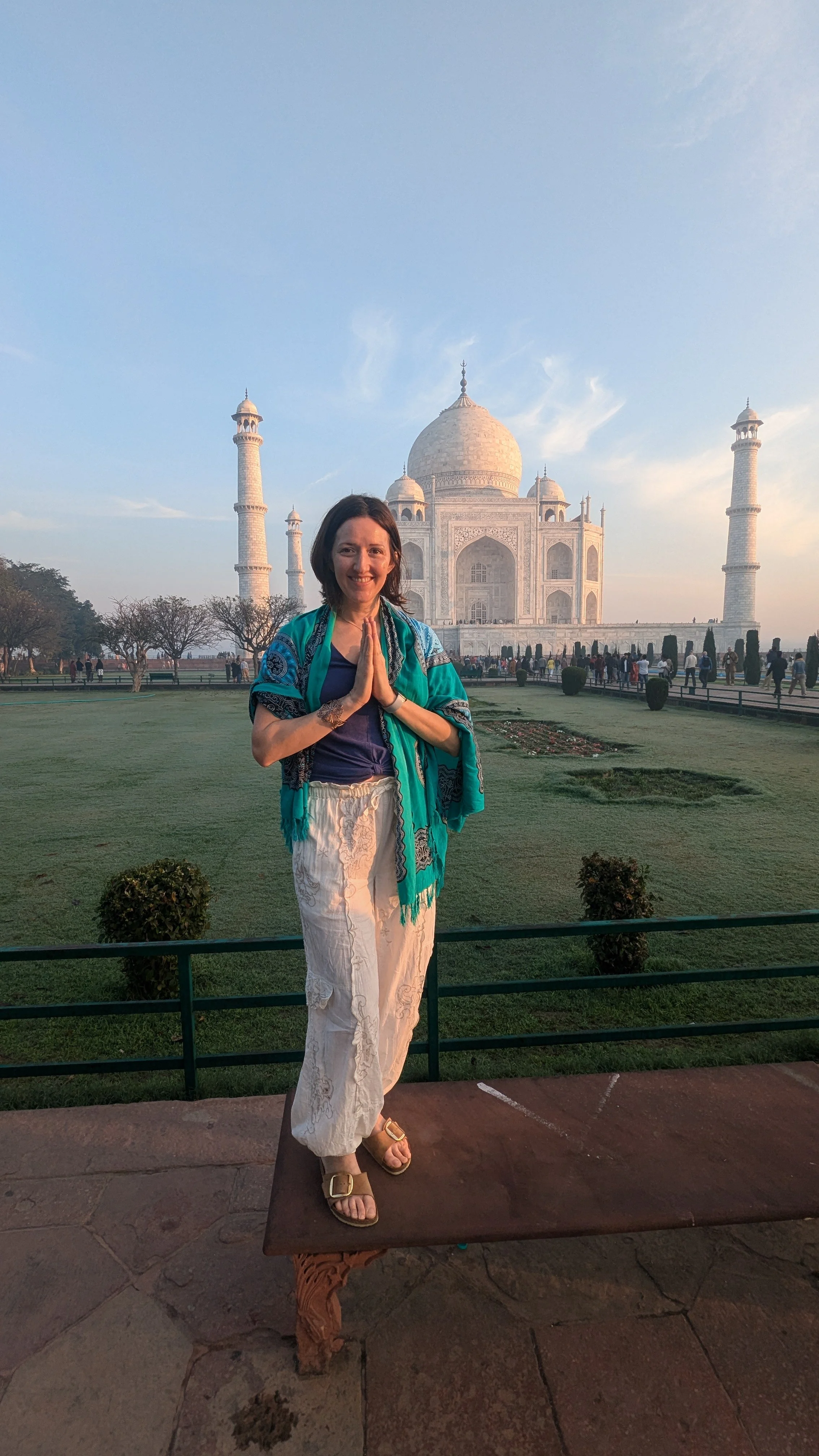 A woman standing on a bench in front of the Taj Mahal with her hands in a prayer position, smiling at the camera during sunset.
