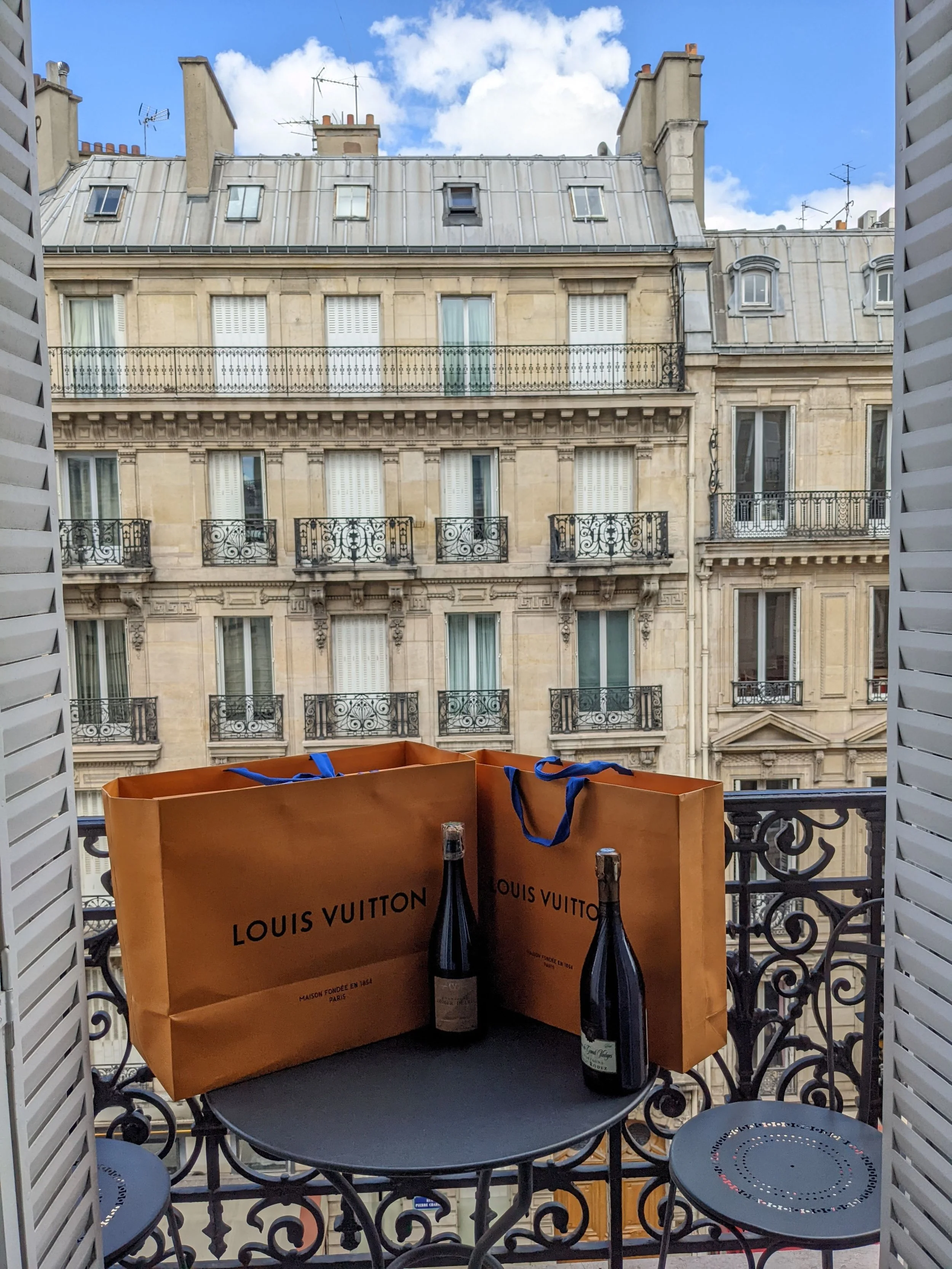 View from a balcony with two Louis Vuitton shopping bags and two bottles of champagne on a small black table, overlooking classic Parisian apartment buildings with ornate balconies and a partly cloudy sky.