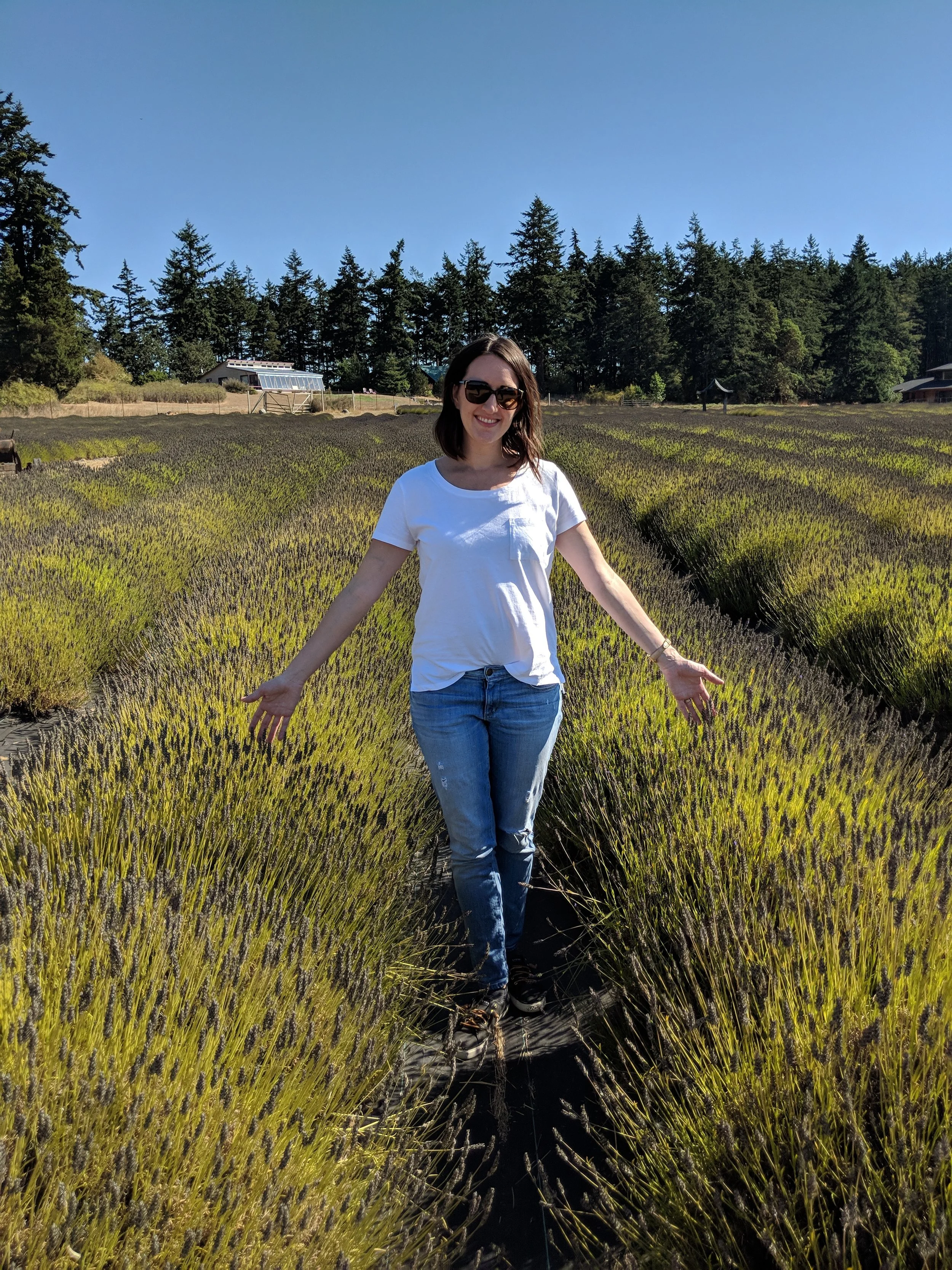 A woman in sunglasses wearing a white t-shirt and jeans standing with arms outstretched in a lavender field under a clear blue sky.