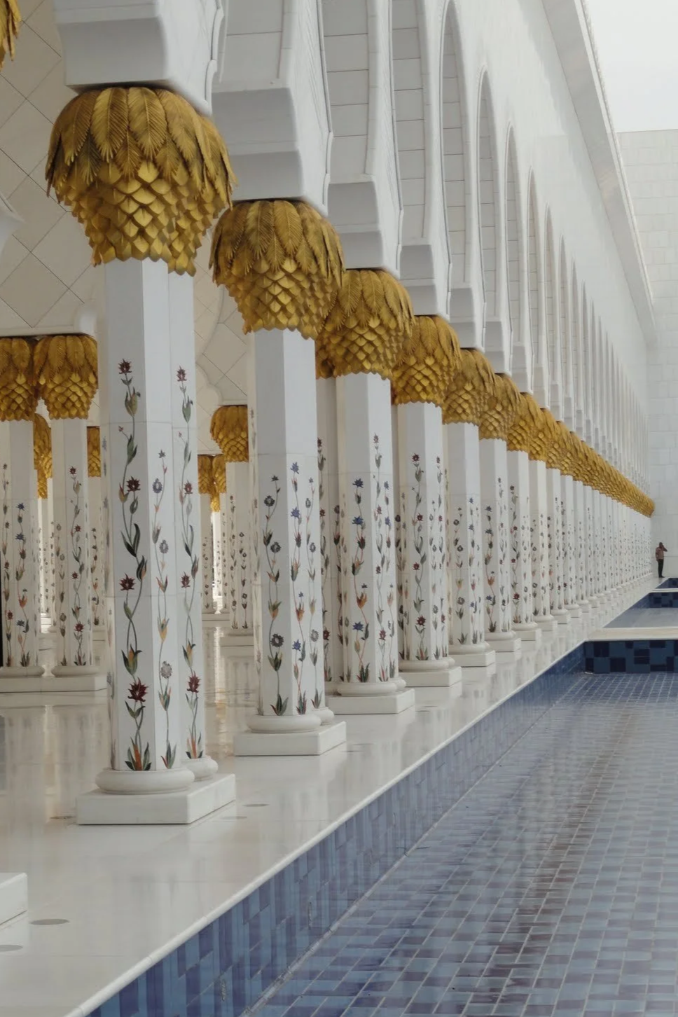 Interior of a building with white columns decorated with colorful floral patterns, topped with golden, leaf-shaped designs, and a blue-tiled pool or fountain on the floor.
