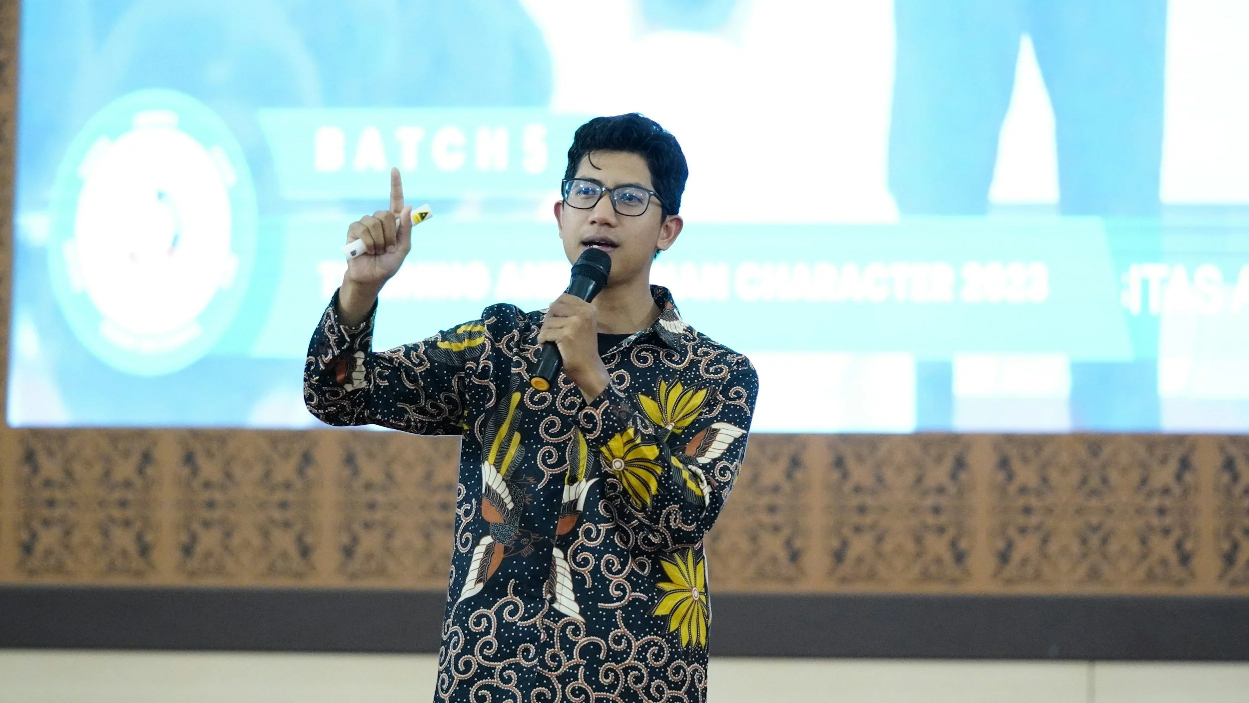 A lived experience key-note speaker wearing glasses, a black and yellow floral batik shirt, is speaking into a microphone and gesturing with his right hand during a presentation or speech.