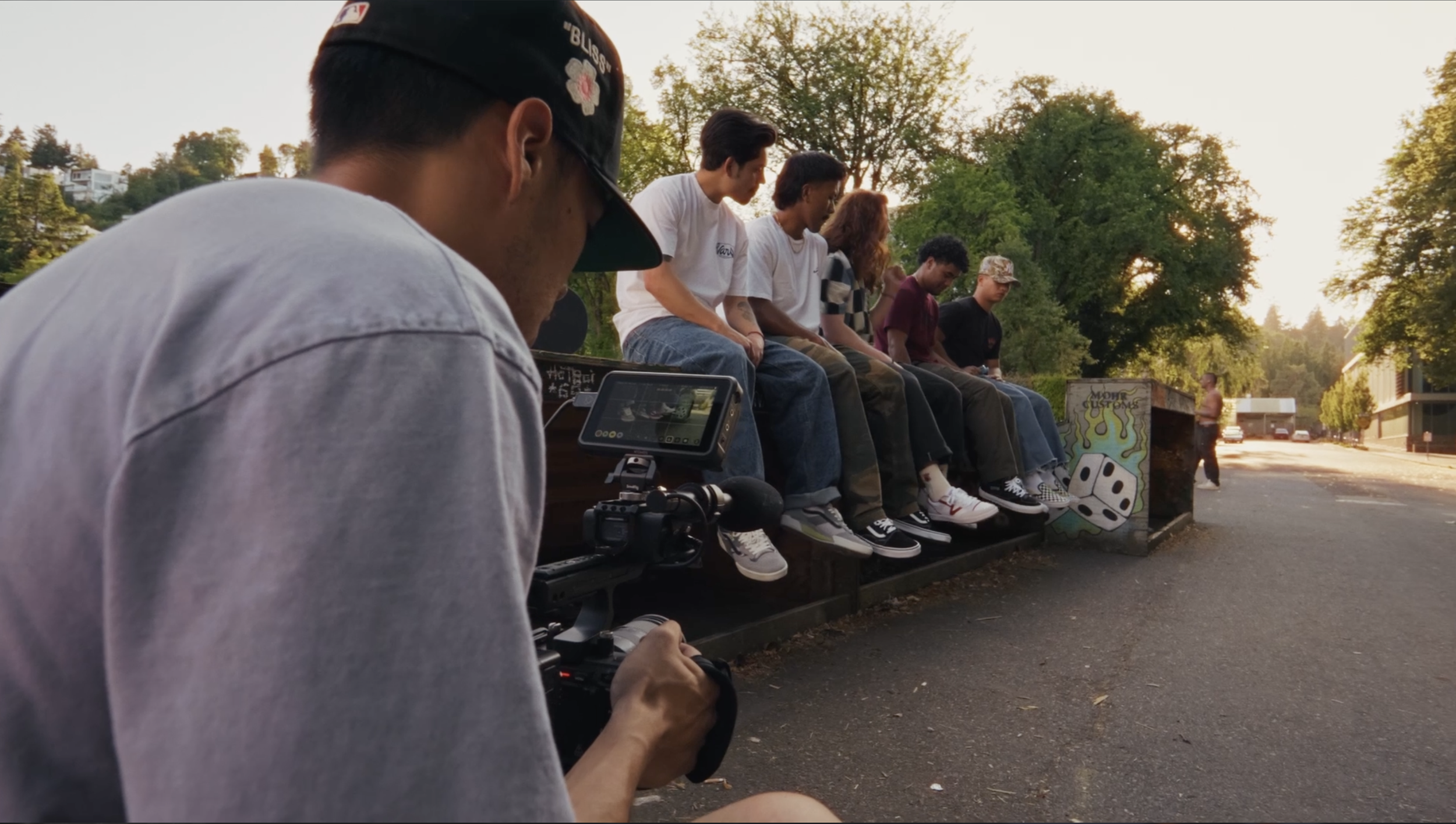 A video producer filming a spec commercial to work on his filmmaking craft. A young man filming a group of people sitting on a bench outdoors during sunset. The group consists of six young people, with trees and buildings in the background.