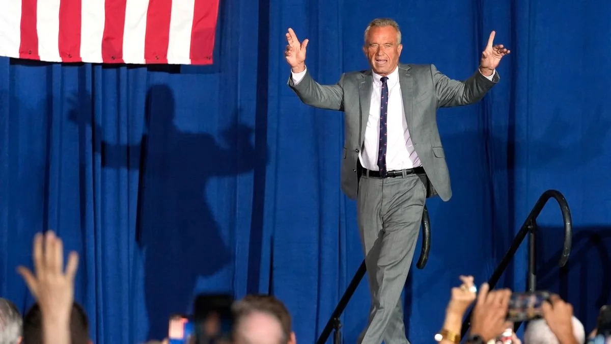 Robert F. Kennedy Jr. in a gray suit on a stage with a blue curtain backdrop, raising his hands in a gesture, in front of an American flag. Audience members are visible in the foreground.