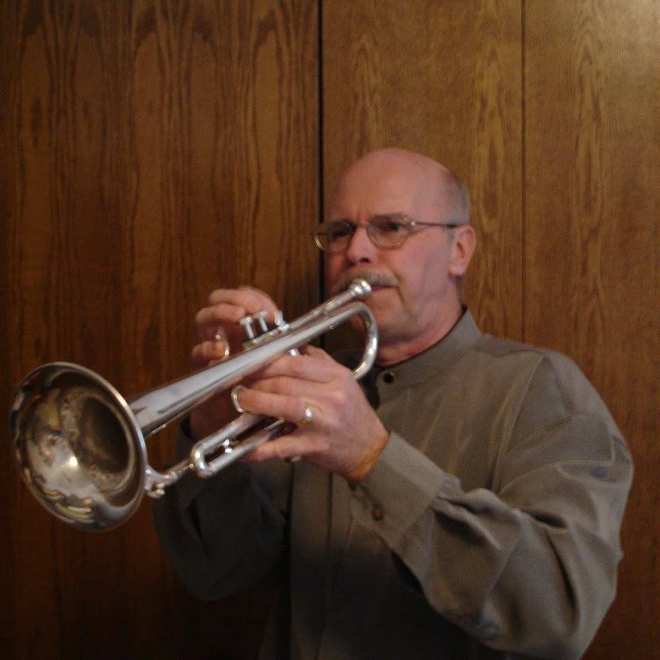 An older man with glasses playing a silver trumpet in front of a wood-paneled wall.