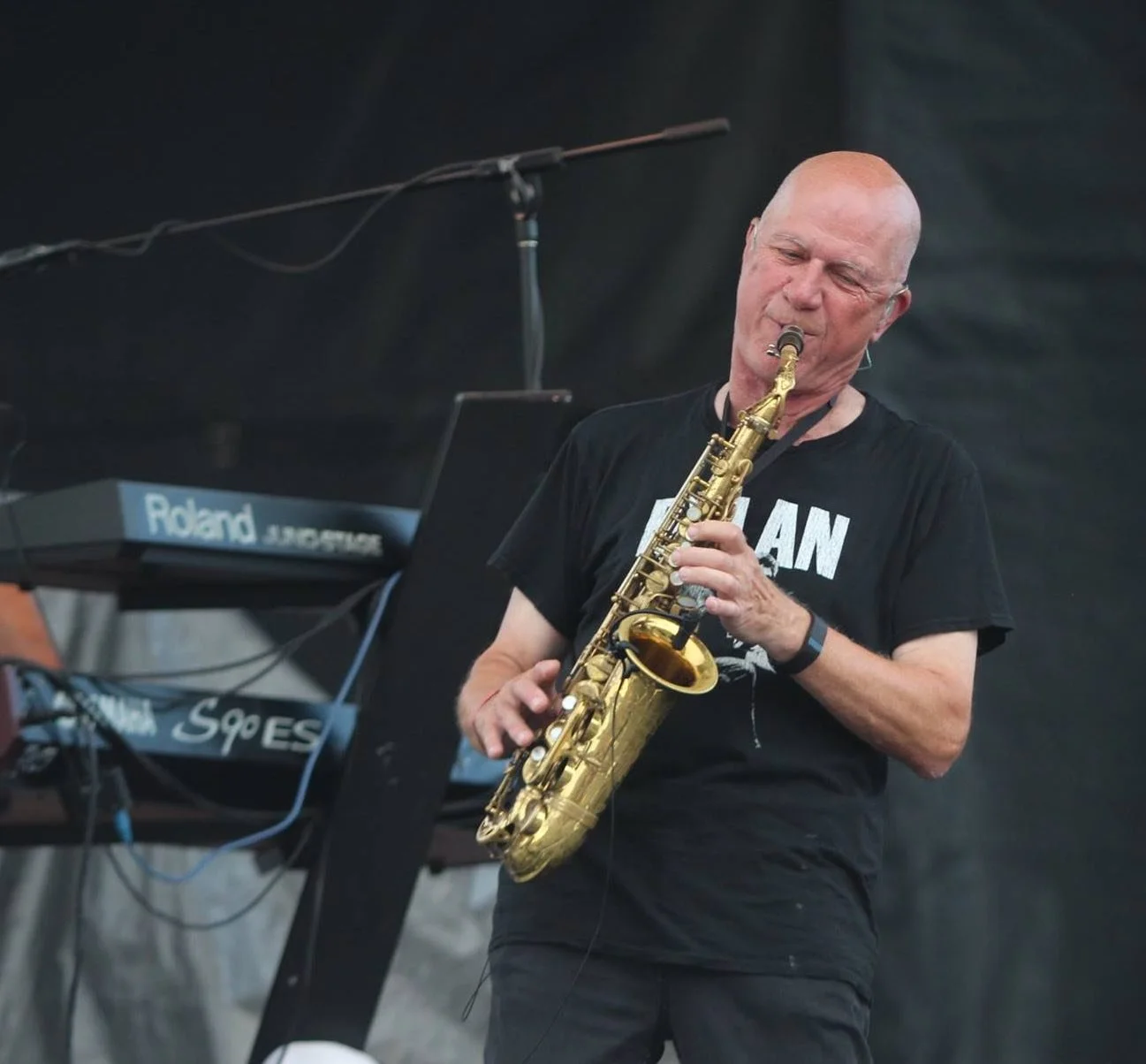 A man playing a gold saxophone during a live music performance, with a keyboard in the background.