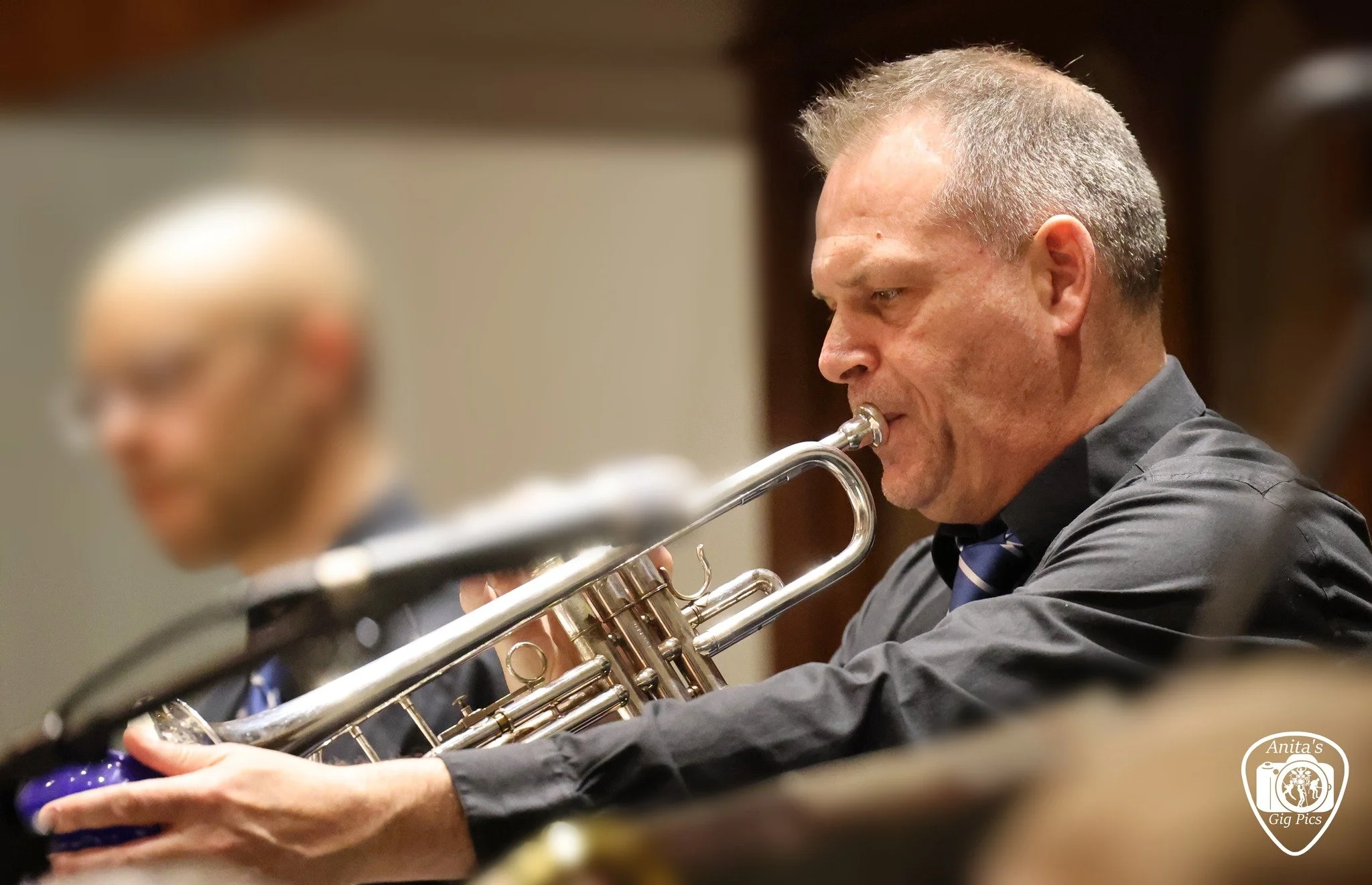 A man playing a trumpet in a concert hall, with another musician slightly out of focus in the background.